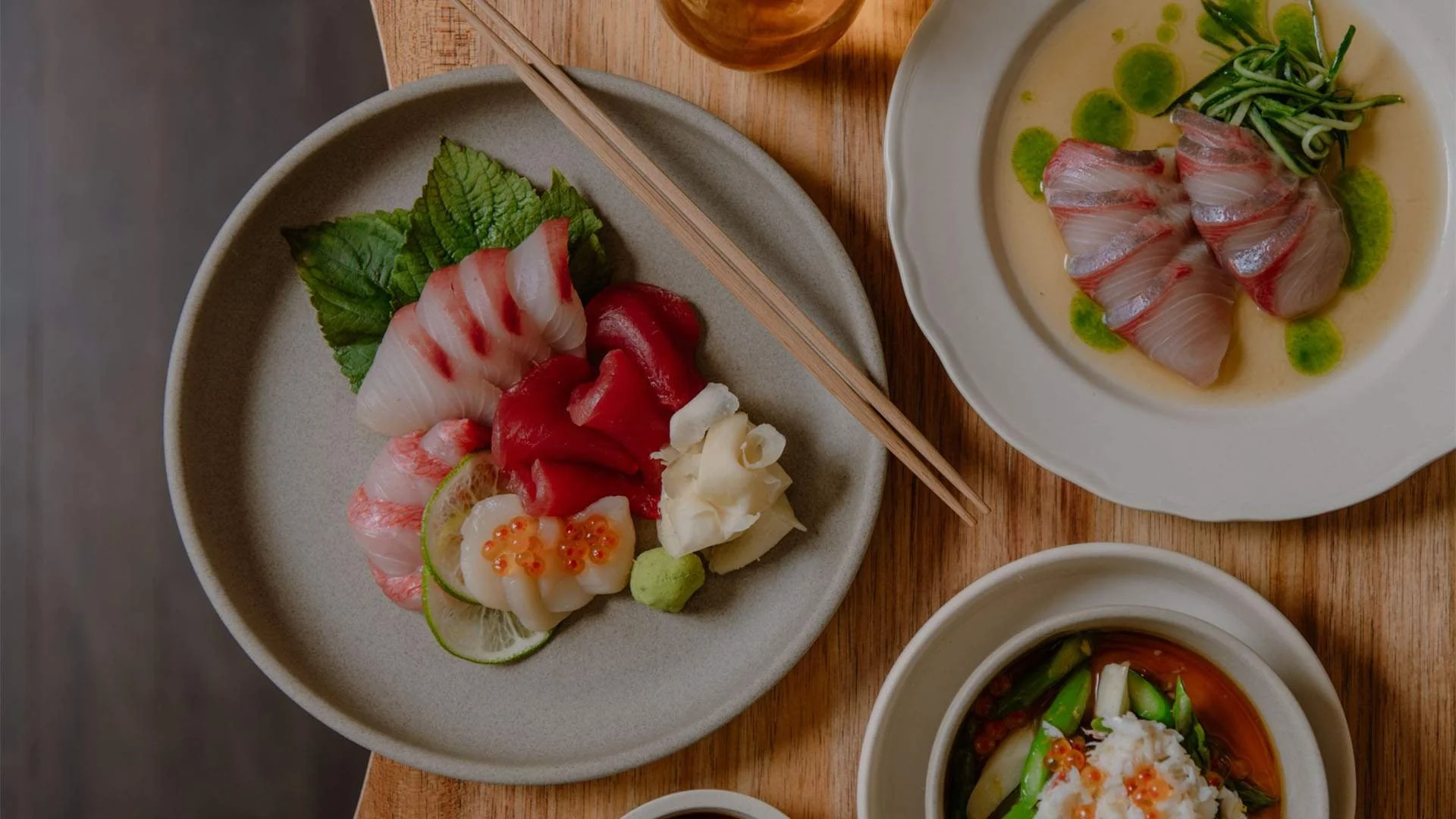Assorted sashimi including tuna, scallop, and white fish with garnishes on a beige plate, and a bowl of sliced raw fish with green sauce, on a wooden table.