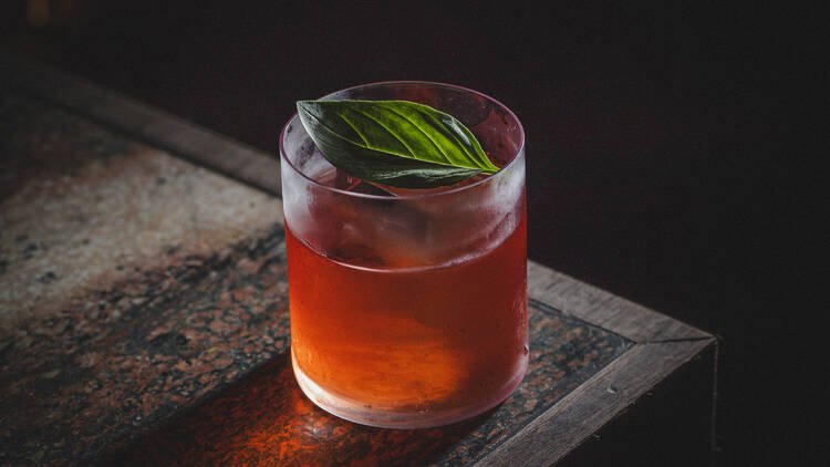 A glass of reddish-colored beverage with ice and a fresh basil leaf on top, placed on a dark wooden surface.