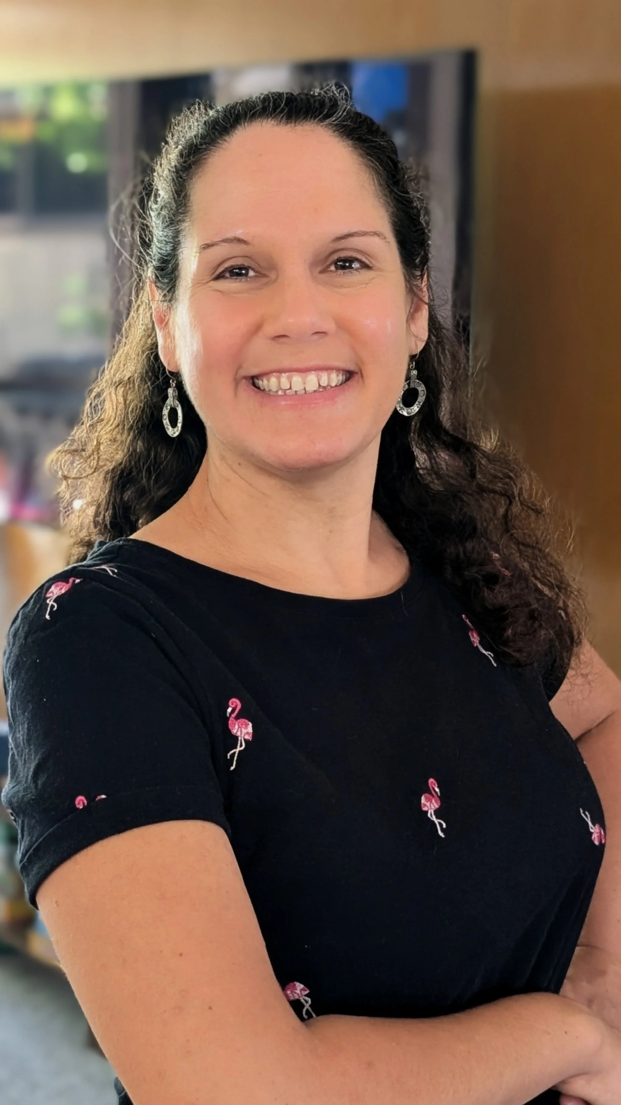 A smiling woman with dark curly hair wearing a black shirt with pink flamingo patterns and silver earrings, standing with her arms crossed indoors.