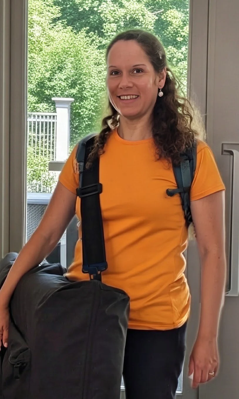 A woman wearing an orange t-shirt, carrying a large black bag, standing inside near a door with a view of green trees outside.