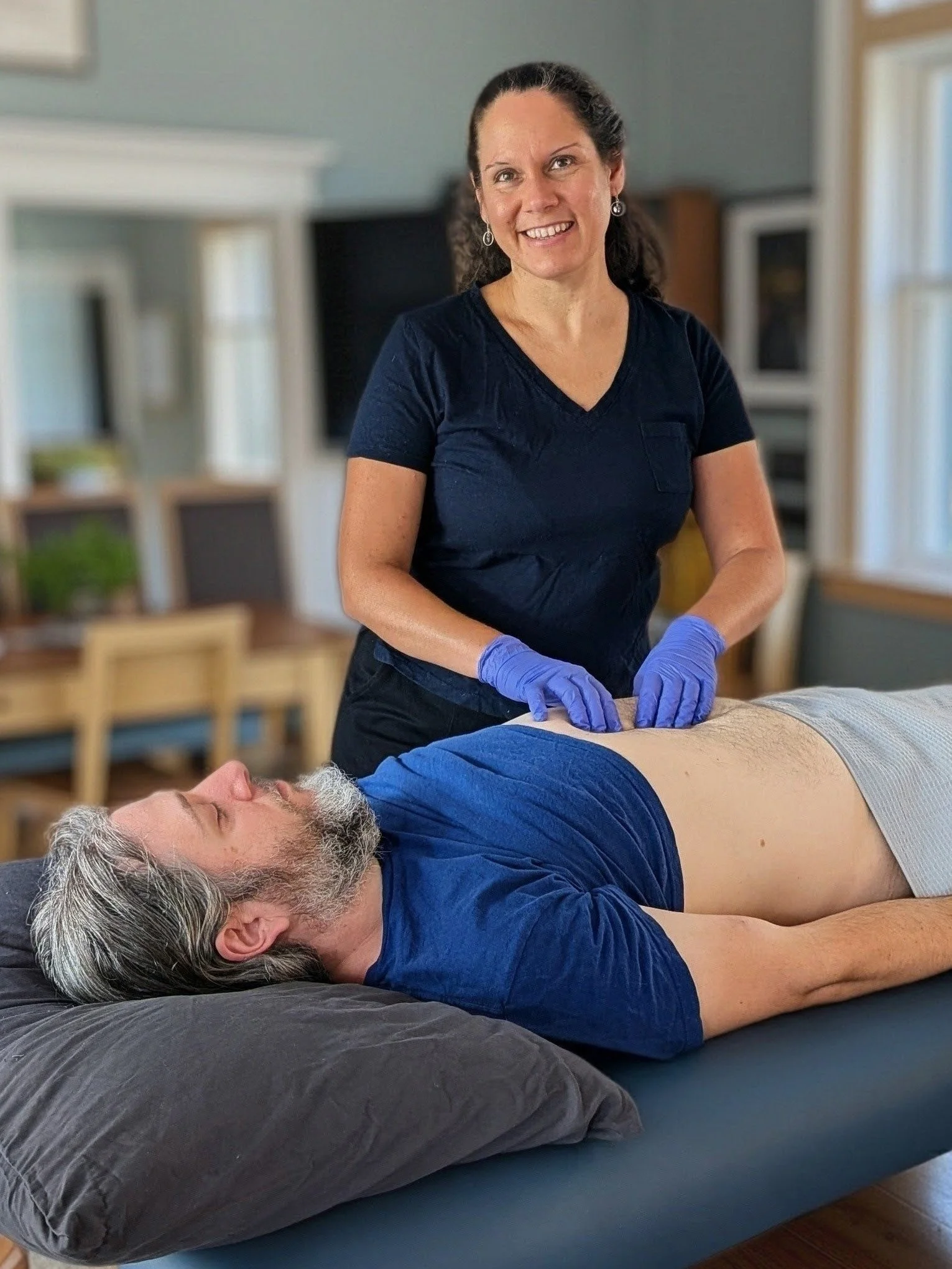 A woman in a black shirt and purple gloves practicing cupping therapy on a man lying face-up on a bed, in a cozy indoor setting with wooden furniture and windows.