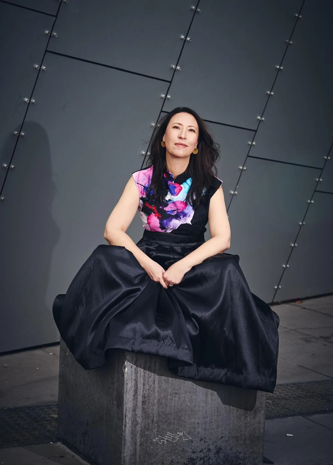 A woman sitting on a concrete block against a modern steel wall with rivets, wearing a colorful sleeveless top and large black skirt, looking at the camera with a slight smile.