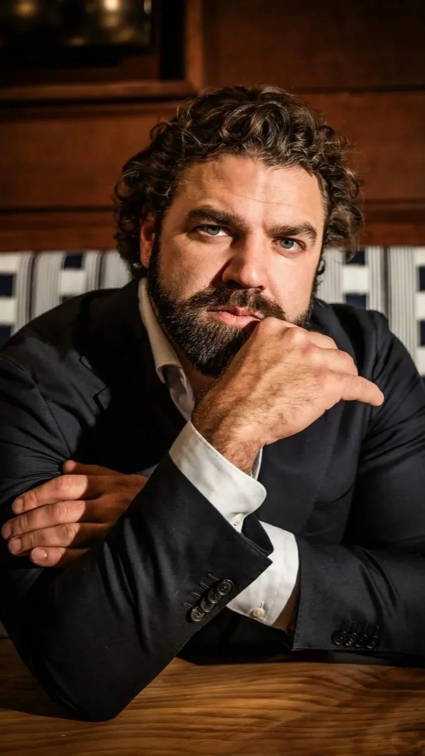 A man with curly hair and a beard sitting at a wooden table, wearing a black suit and white shirt, with his chin resting on his hand, looking thoughtful.