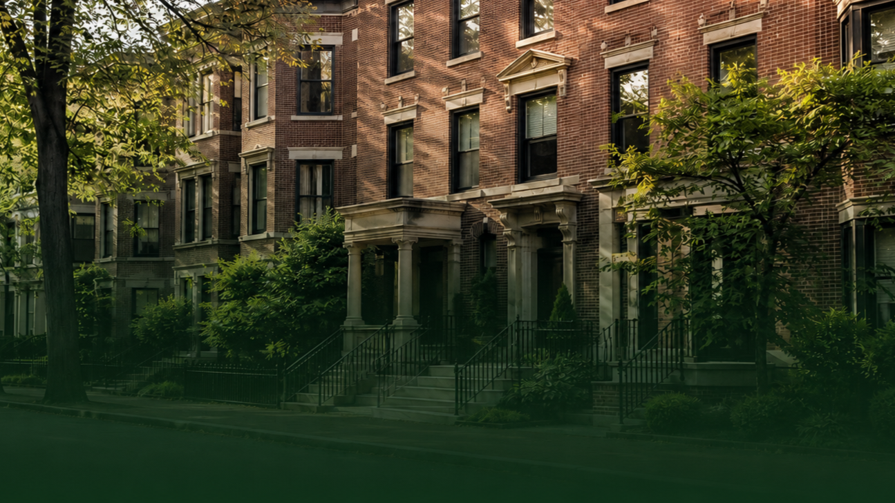 A row of historic brick townhouses with steps leading to porches, surrounded by trees and greenery in an urban neighborhood.