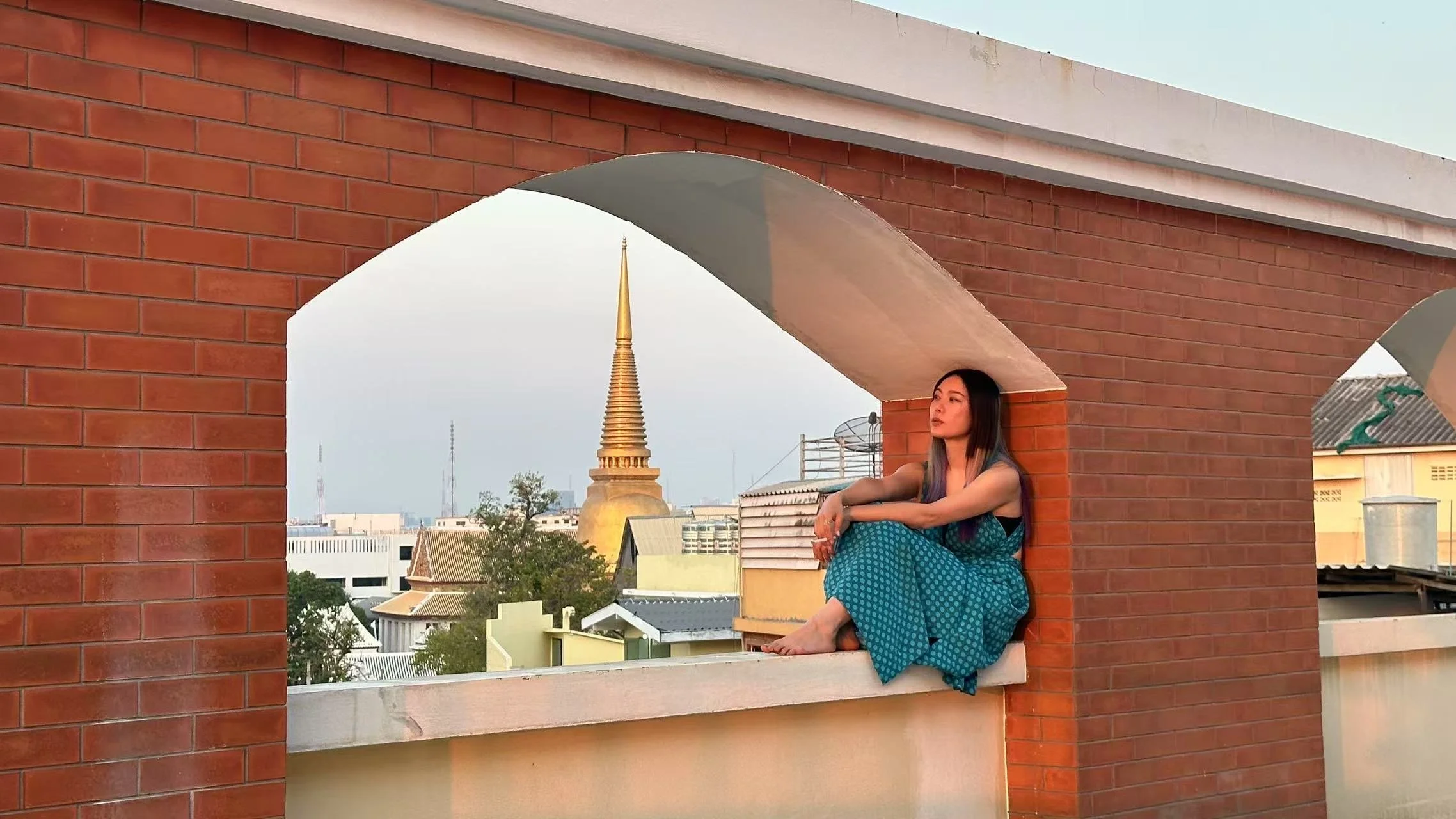 A woman with long dark hair sitting on a ledge on a brick rooftop, with a cityscape and a golden temple spire in the background.