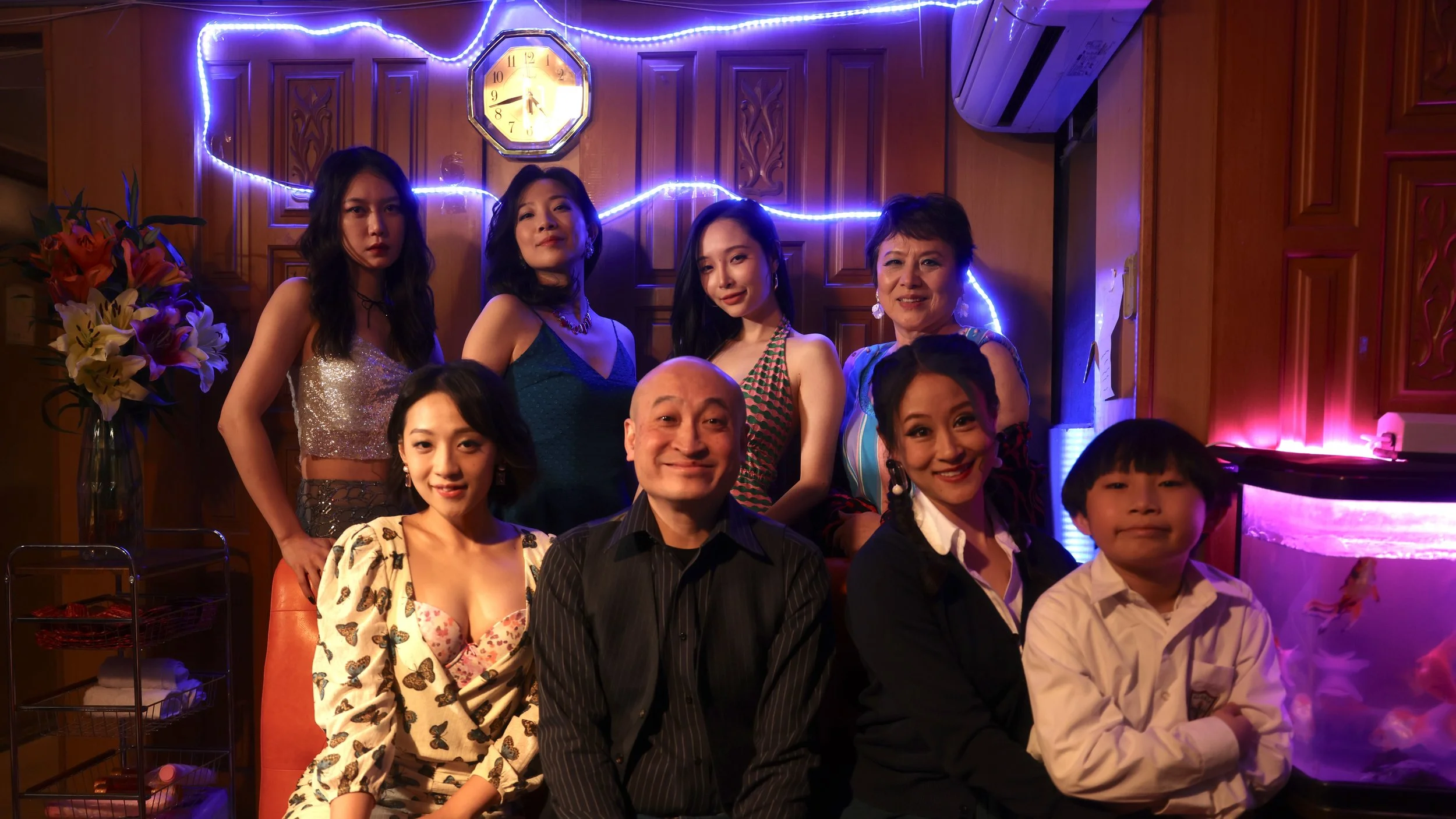 Group of people, mostly women and one young boy, posing for a photo in a warmly lit room decorated with purple lights and a clock on the wall. Some are dressed in colorful, stylish clothing, and a vase of flowers is visible on the left side.