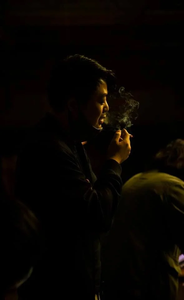 A person in a dark setting is holding a cigarette close to their mouth, with smoke rising from it. The person is in profile, with short dark hair, and appears to be smiling or smiling mildly.