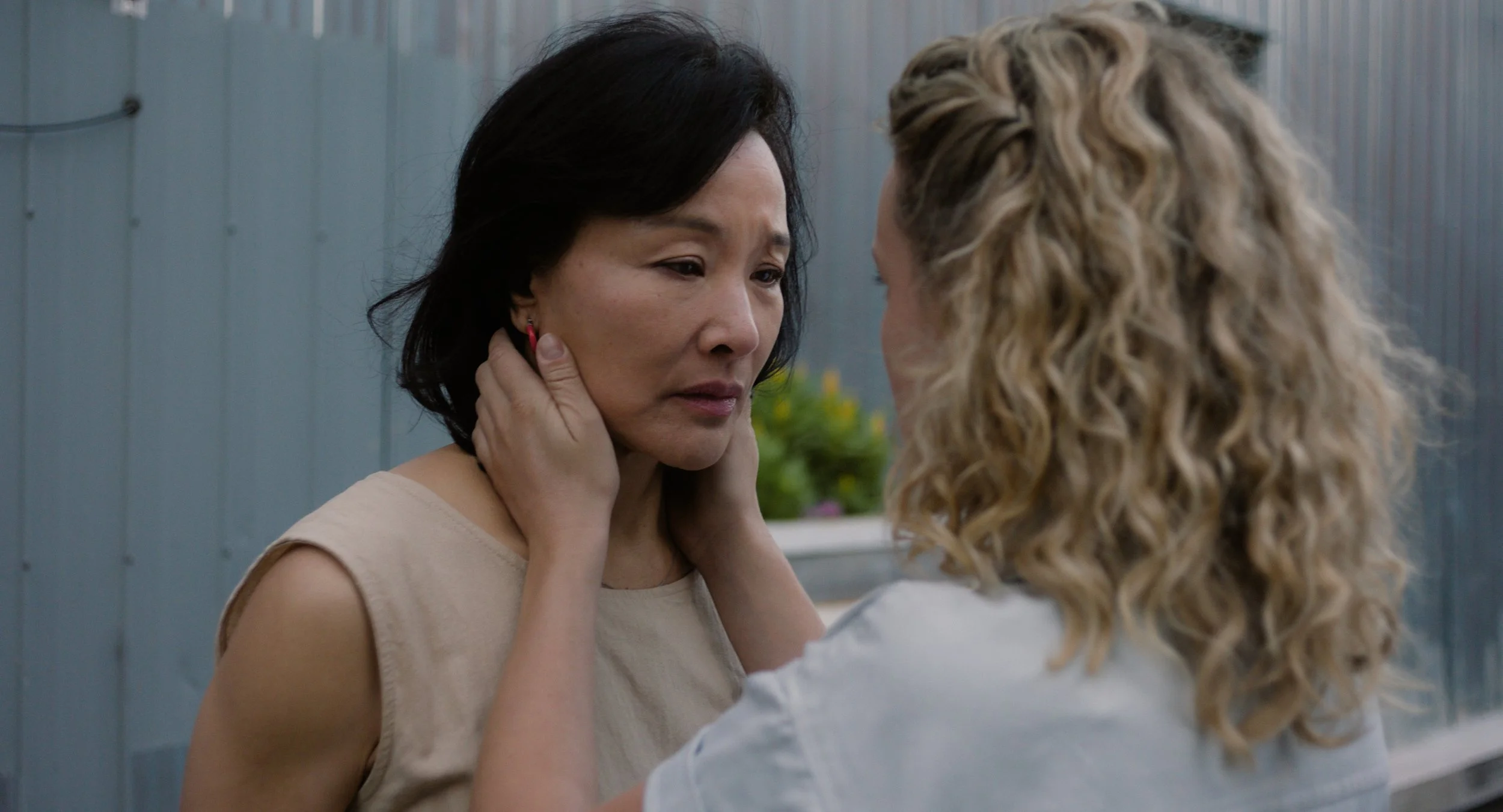 Two women are facing each other closely, with one woman gently holding the other's face. The woman with black hair looks emotional, while the woman with curly blonde hair appears comforting. They are outside, near a blue fence with some greenery and 