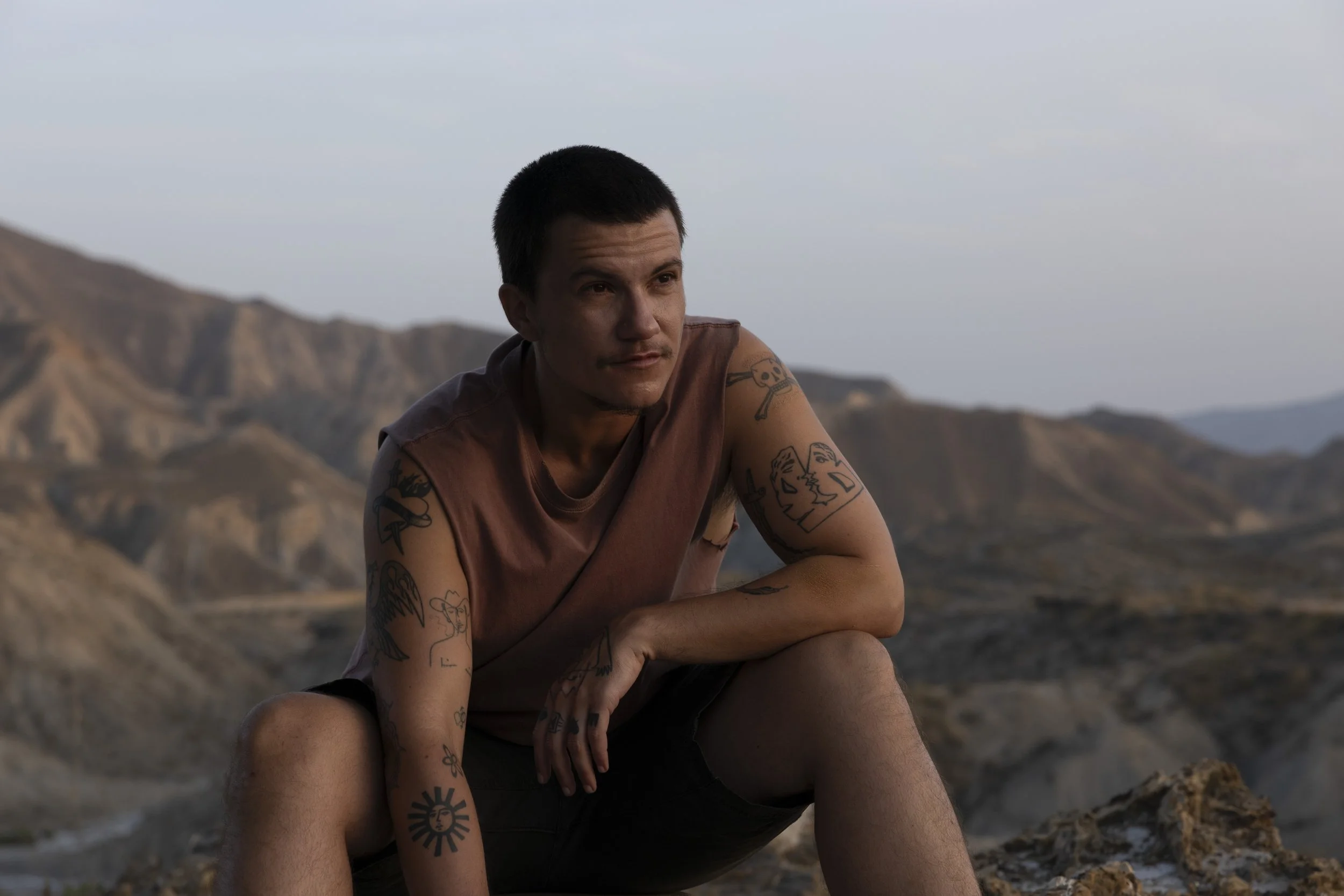 A young man with short black hair and tattoos on his arms, sitting outdoors in a desert landscape with mountains in the background, during the daytime.