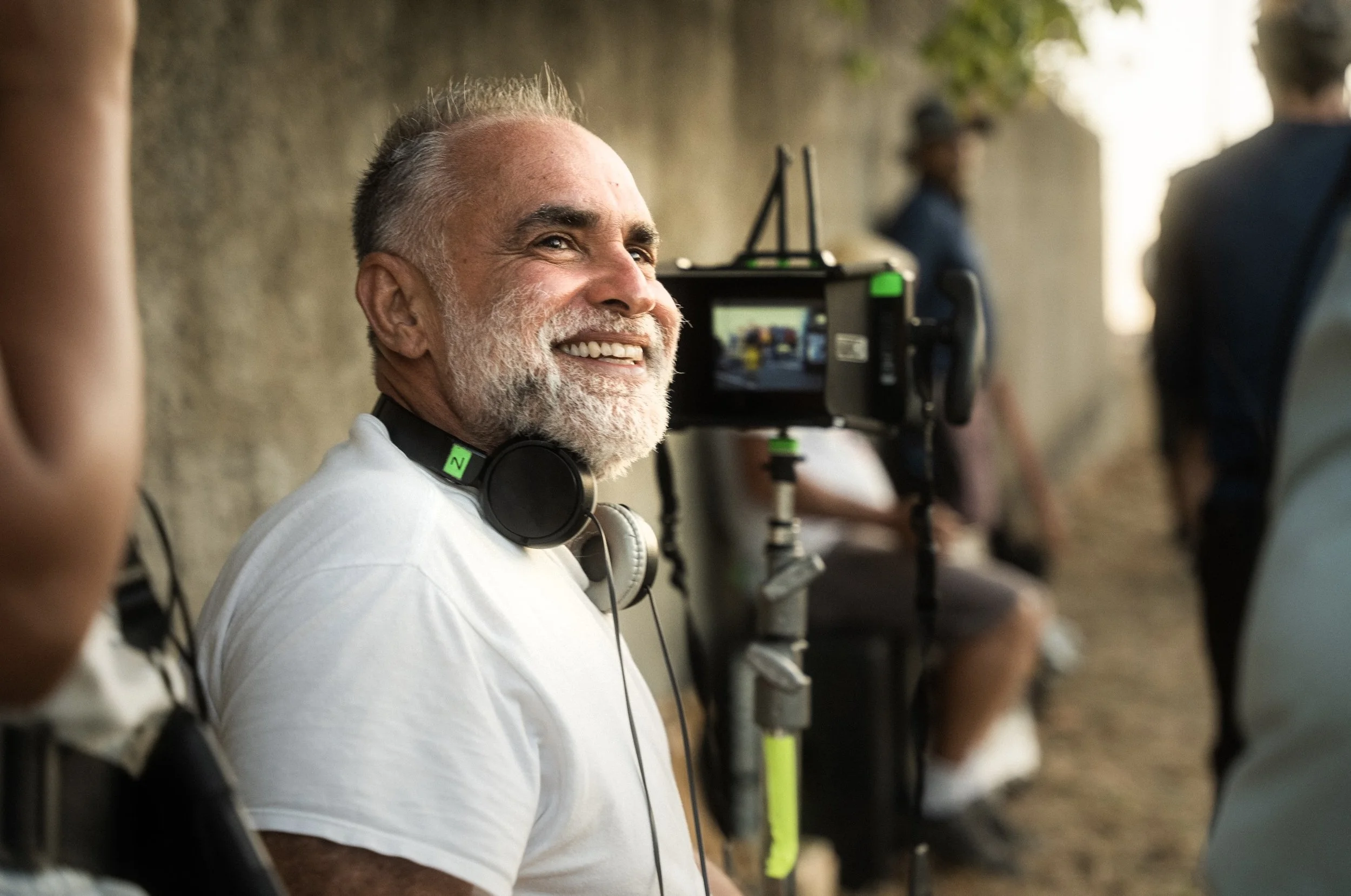A smiling older man with gray hair and a beard wearing headphones around his neck, on a film set with cameras and crew members in the background.