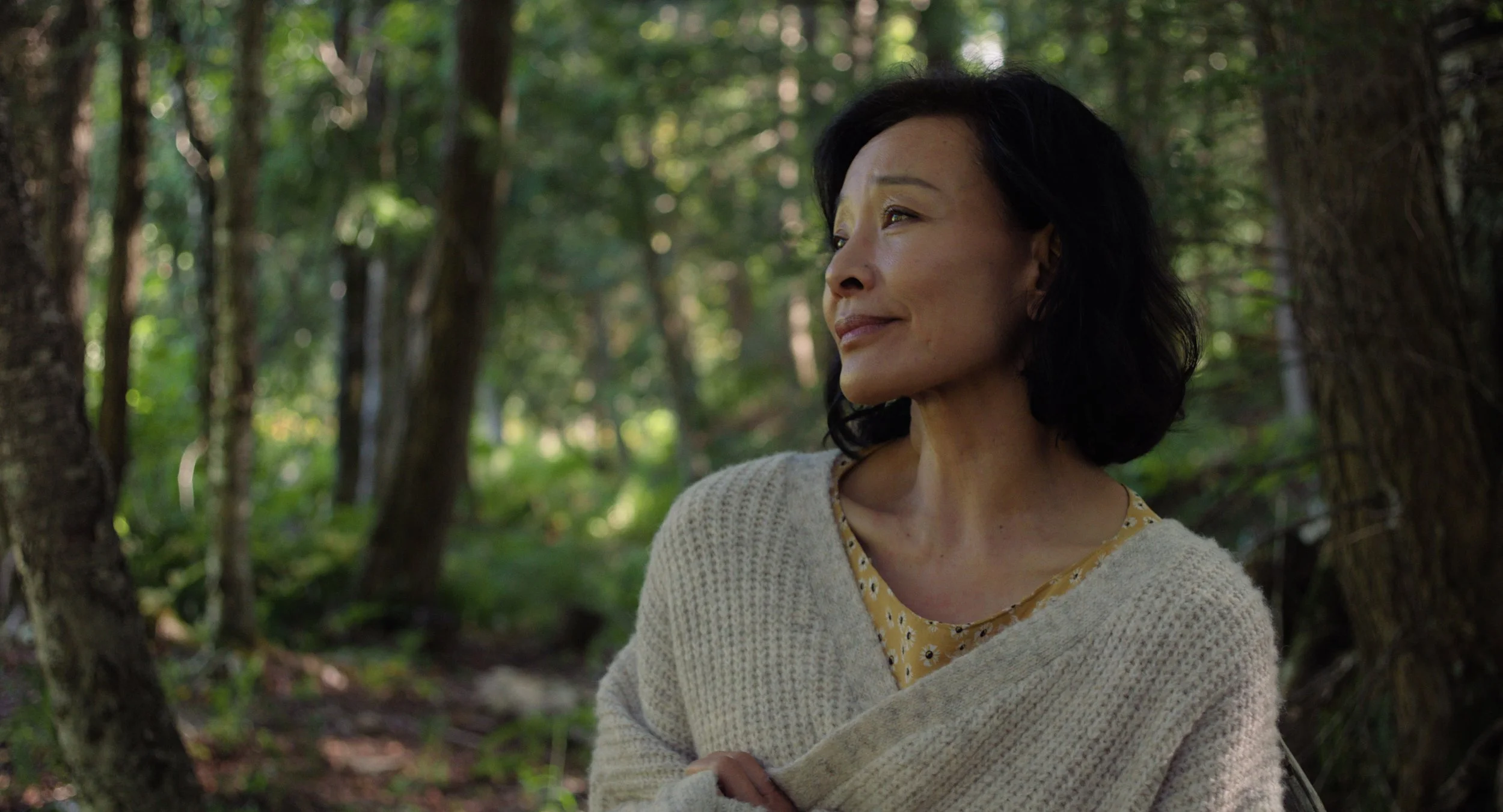 A woman with dark hair and a gentle smile looking to her right, standing in a lush green, wooded outdoor setting with trees and sunlight filtering through the leaves.