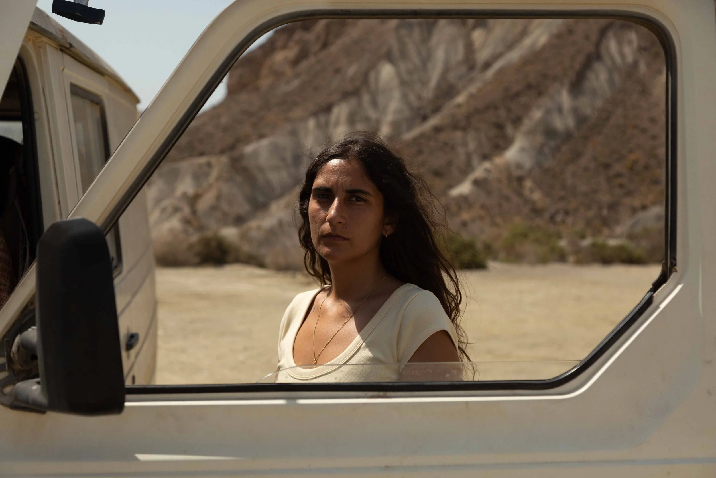 A woman with long wavy dark hair looks out from the side mirror of a white vehicle, with a rocky desert landscape in the background.