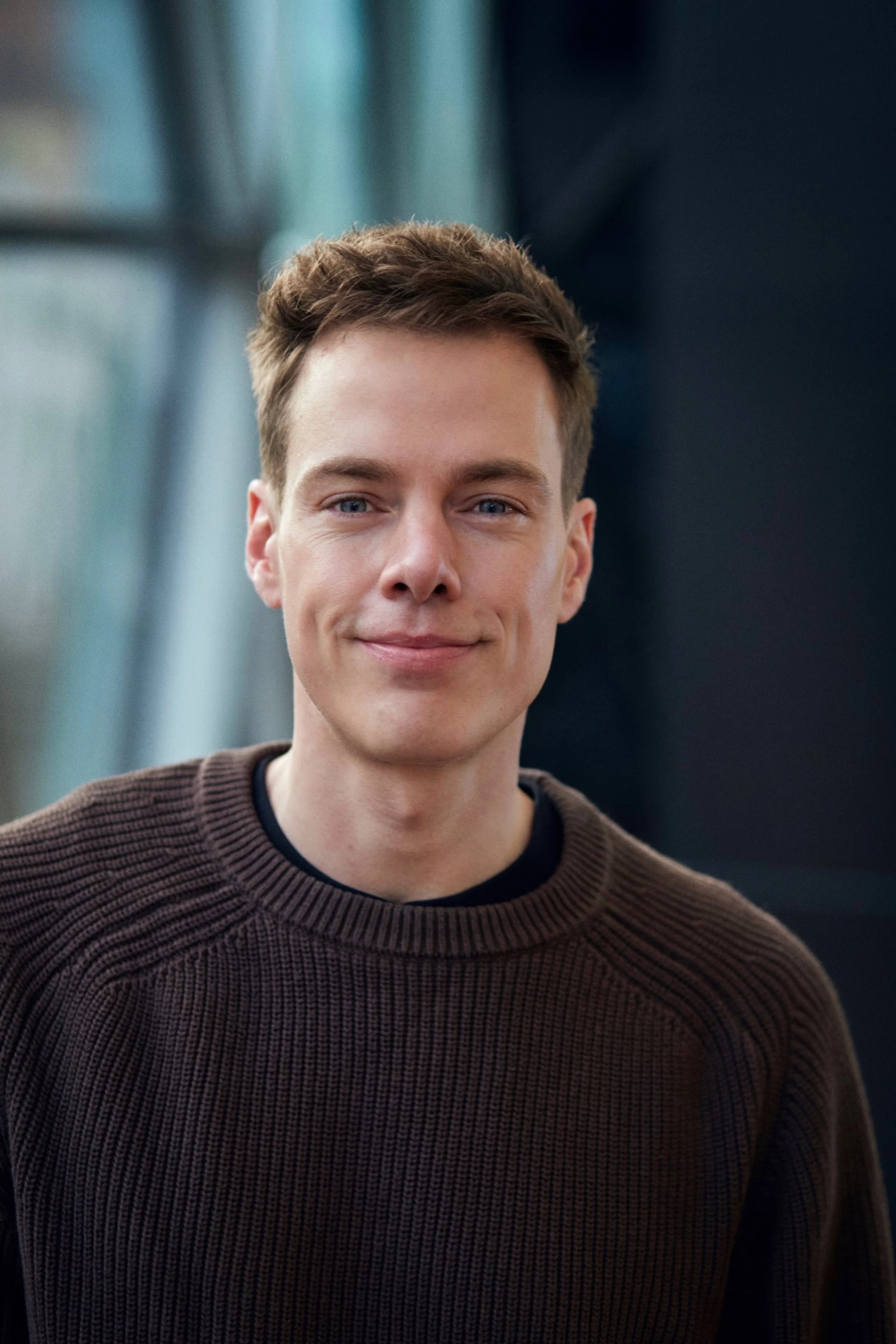 A young man with short, light brown hair and blue eyes smiling at the camera, wearing a dark brown sweater with a ribbed pattern, indoors with a blurred background.