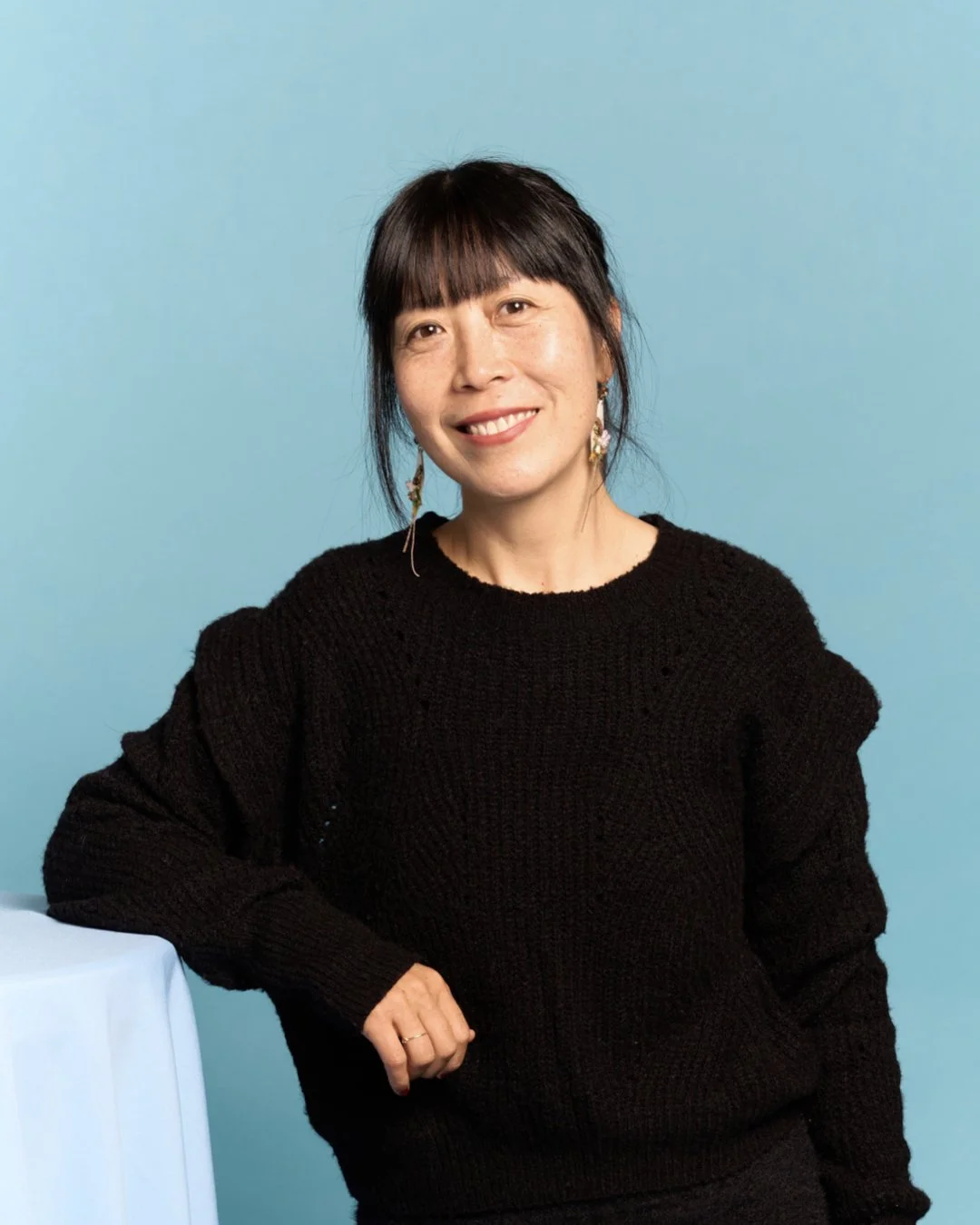 A woman with dark hair and bangs, wearing a black sweater and earrings, smiling while leaning on a table against a light blue background.