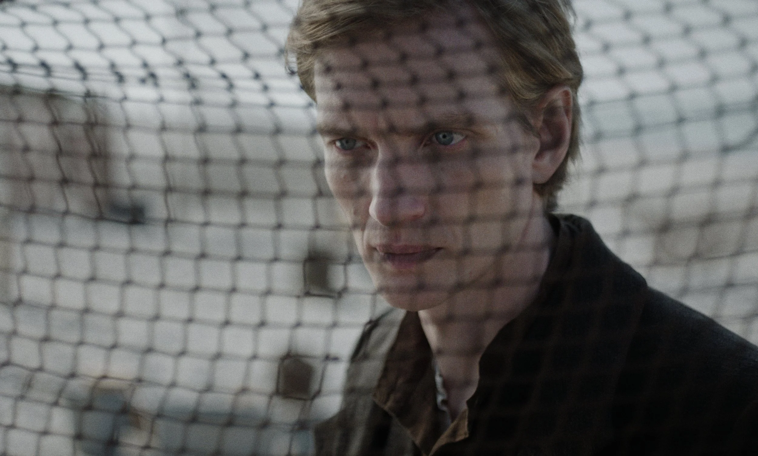 A young man with blonde hair and blue eyes looking through a chain-link fence.