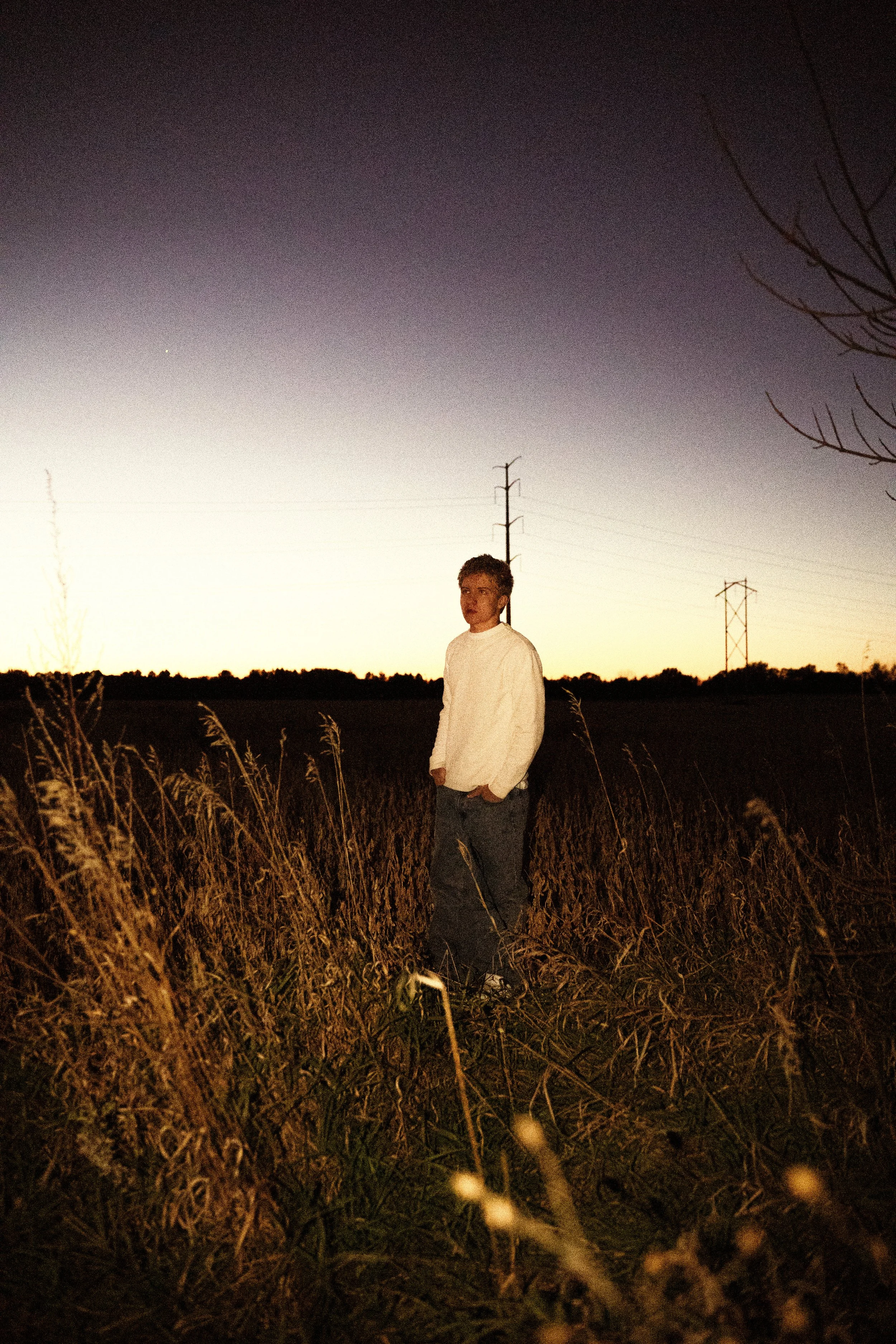 A young man standing in a field of tall grass during dusk or early evening, with power lines and trees visible in the background.