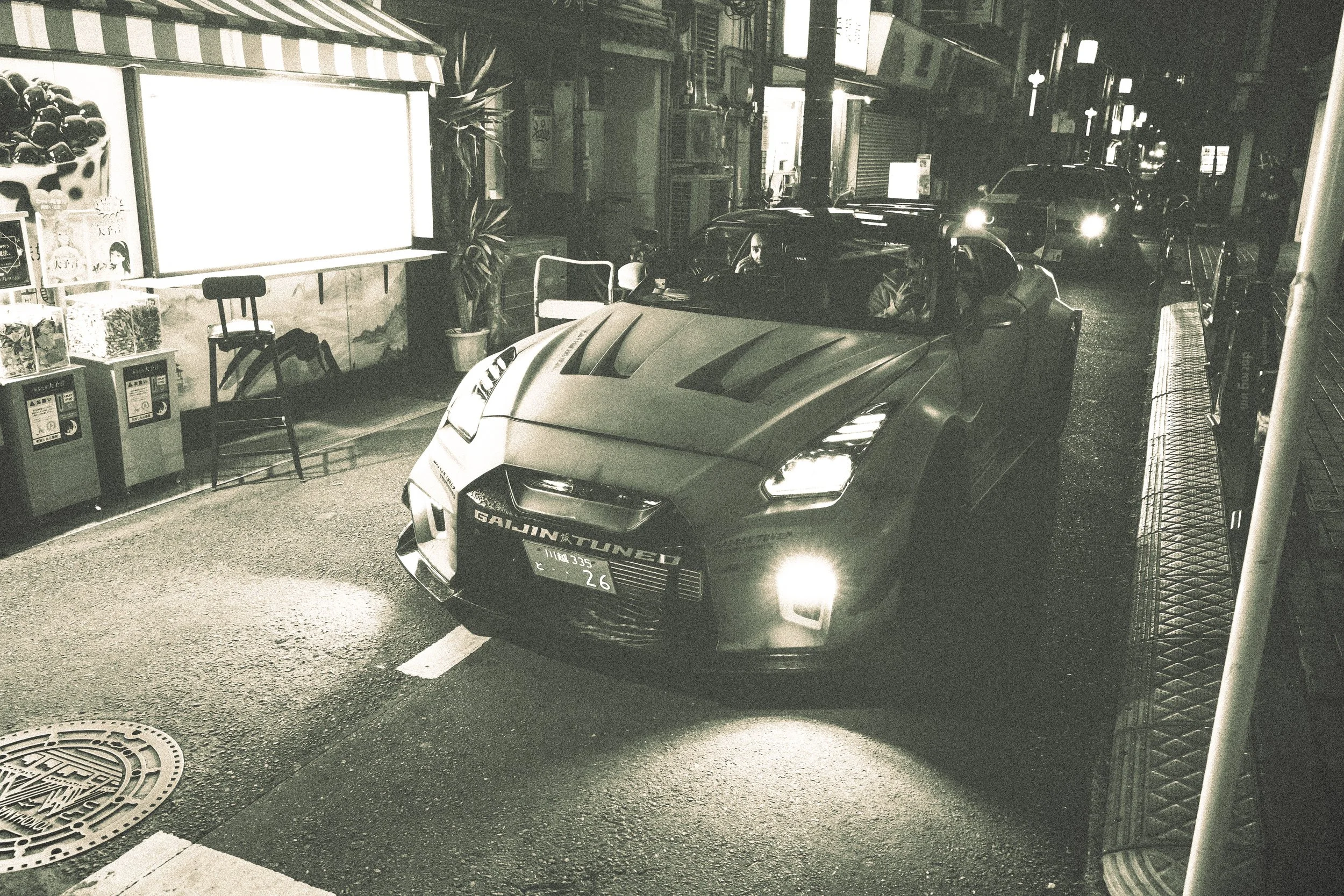 Nighttime street scene with a sports car in front of a small storefront, illuminated signs, and parked cars along the road.