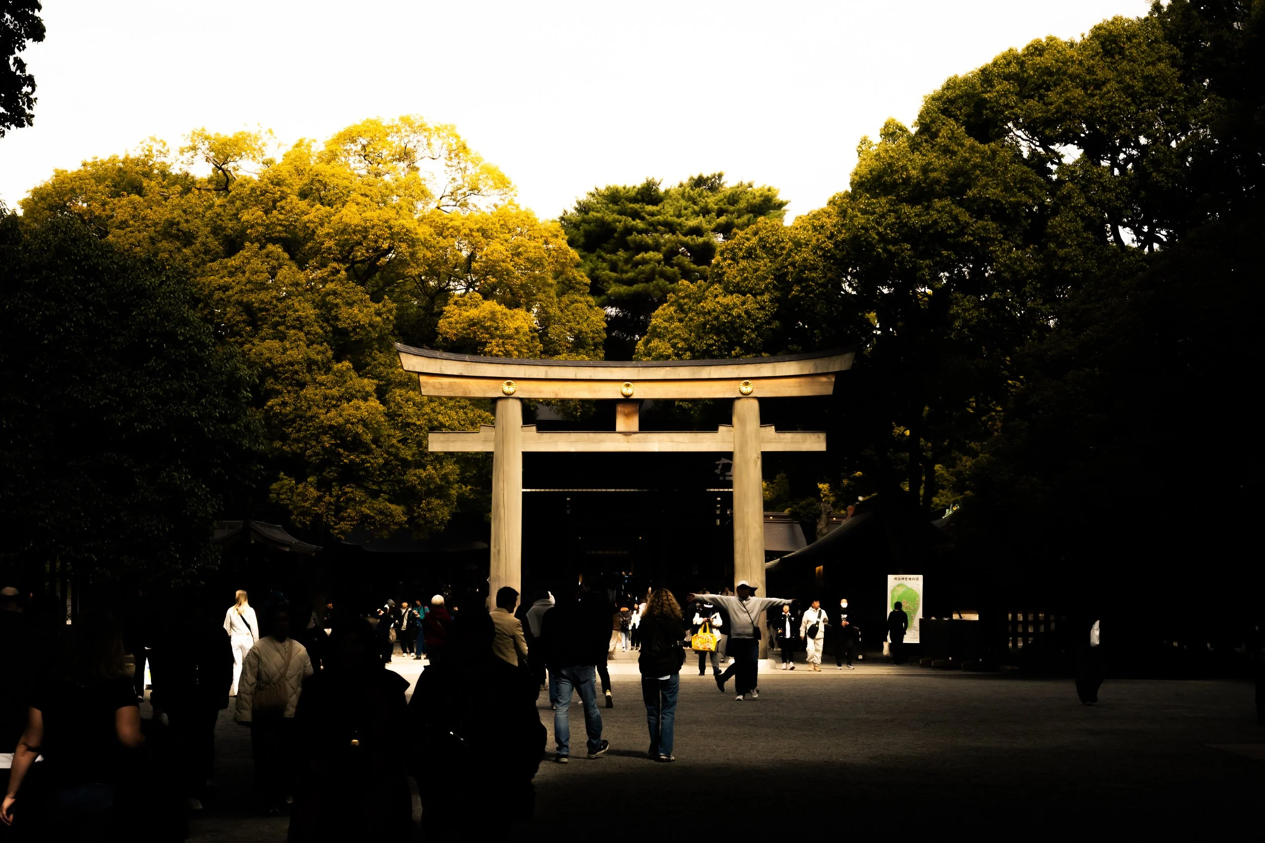 Traditional Japanese torii gate surrounded by green trees with people walking underneath in a park or shrine setting.