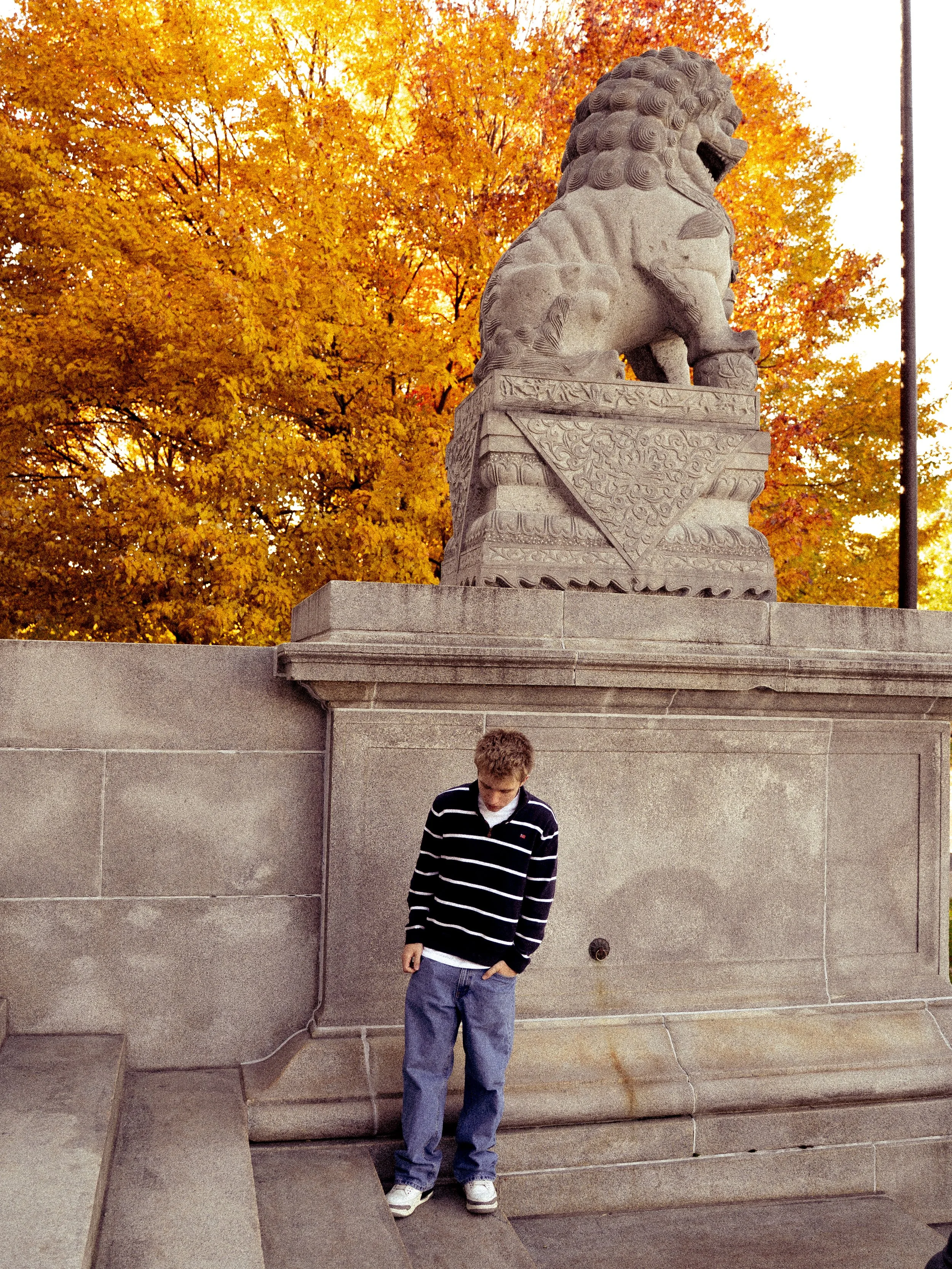 A young man with blond hair, wearing a black and white striped shirt and blue jeans, standing in front of a stone wall and monument with a carved stone lion statue on top. The background is filled with orange and yellow autumn trees.