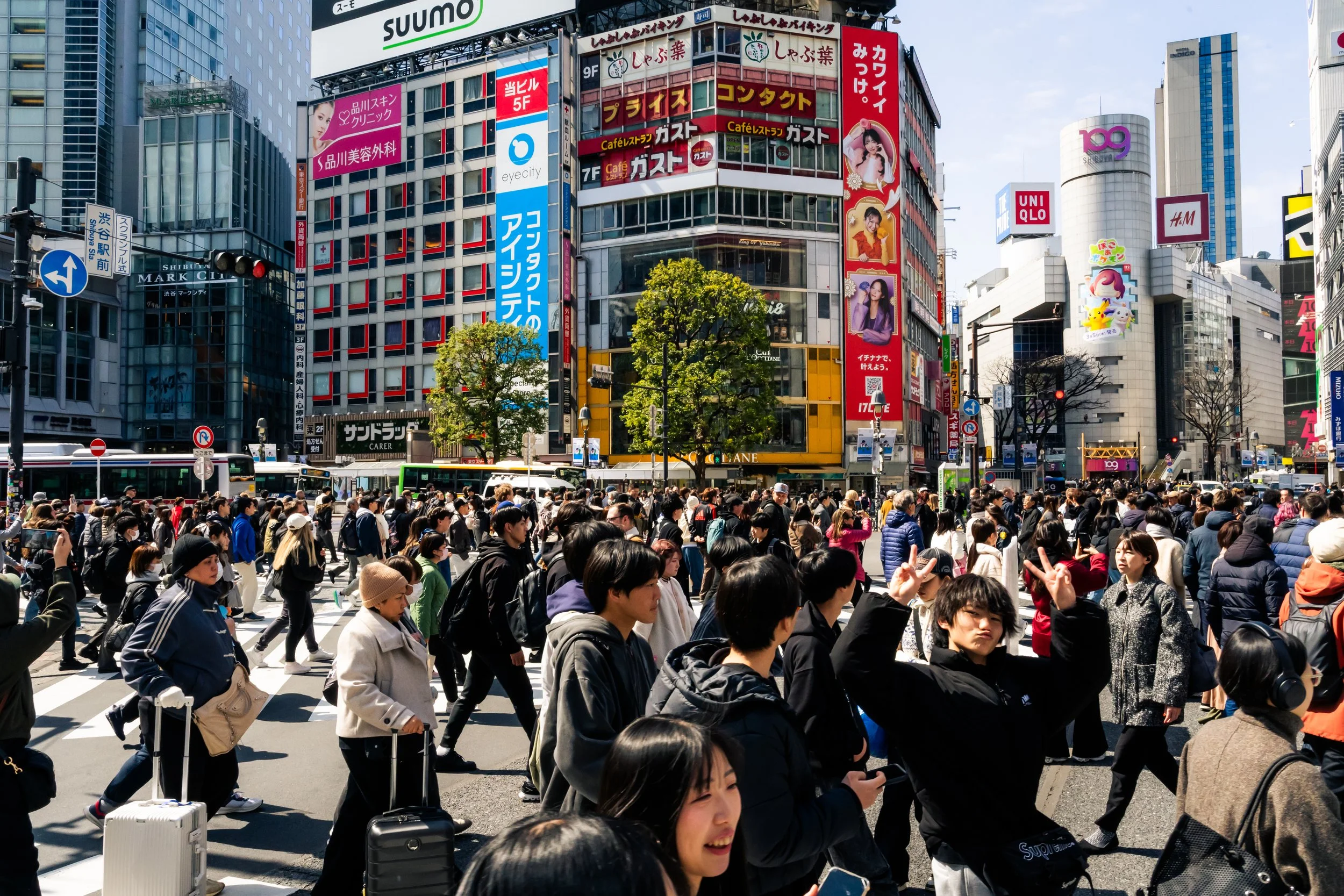 Crowded city street with many pedestrians crossing at a crosswalk, surrounded by tall buildings with colorful advertisements and signs in Japanese and English.
