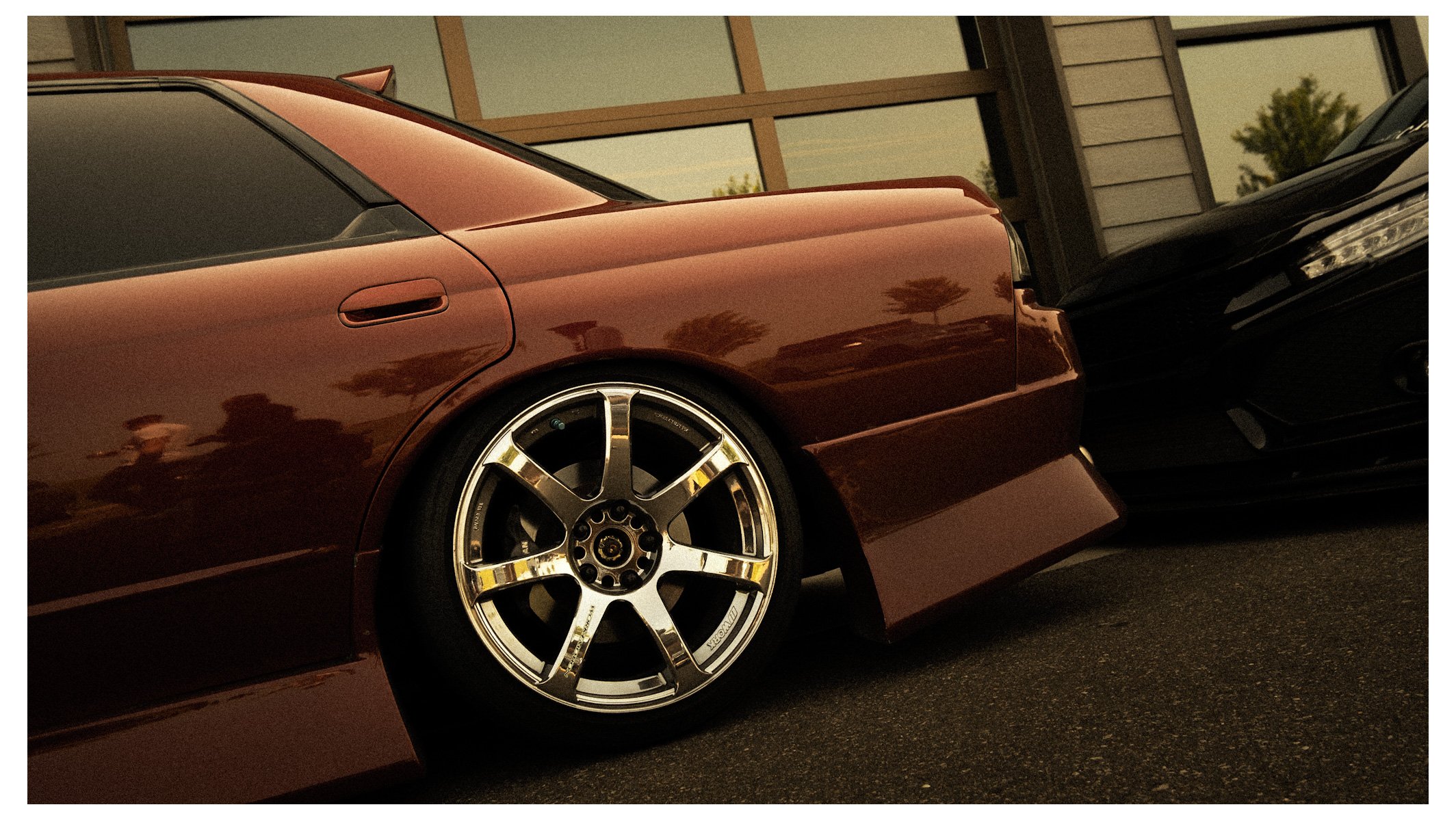 Close-up of a maroon sports car with a chrome rim wheel parked outdoors near a modern building with glass windows and a tree in the background.