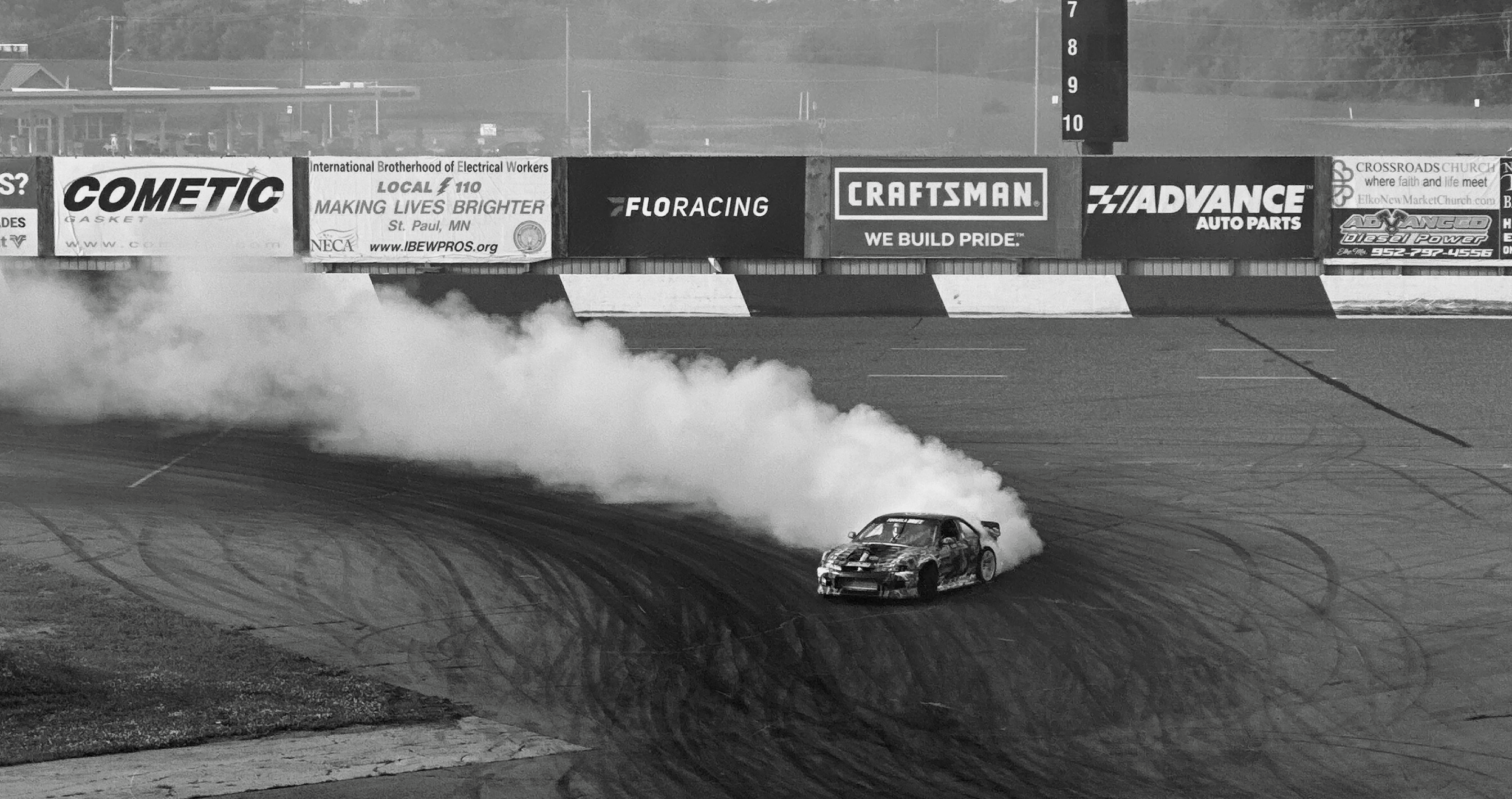 A race car performing a drift on a racetrack with smoke coming from the tires, with banners advertising various auto parts brands in the background.