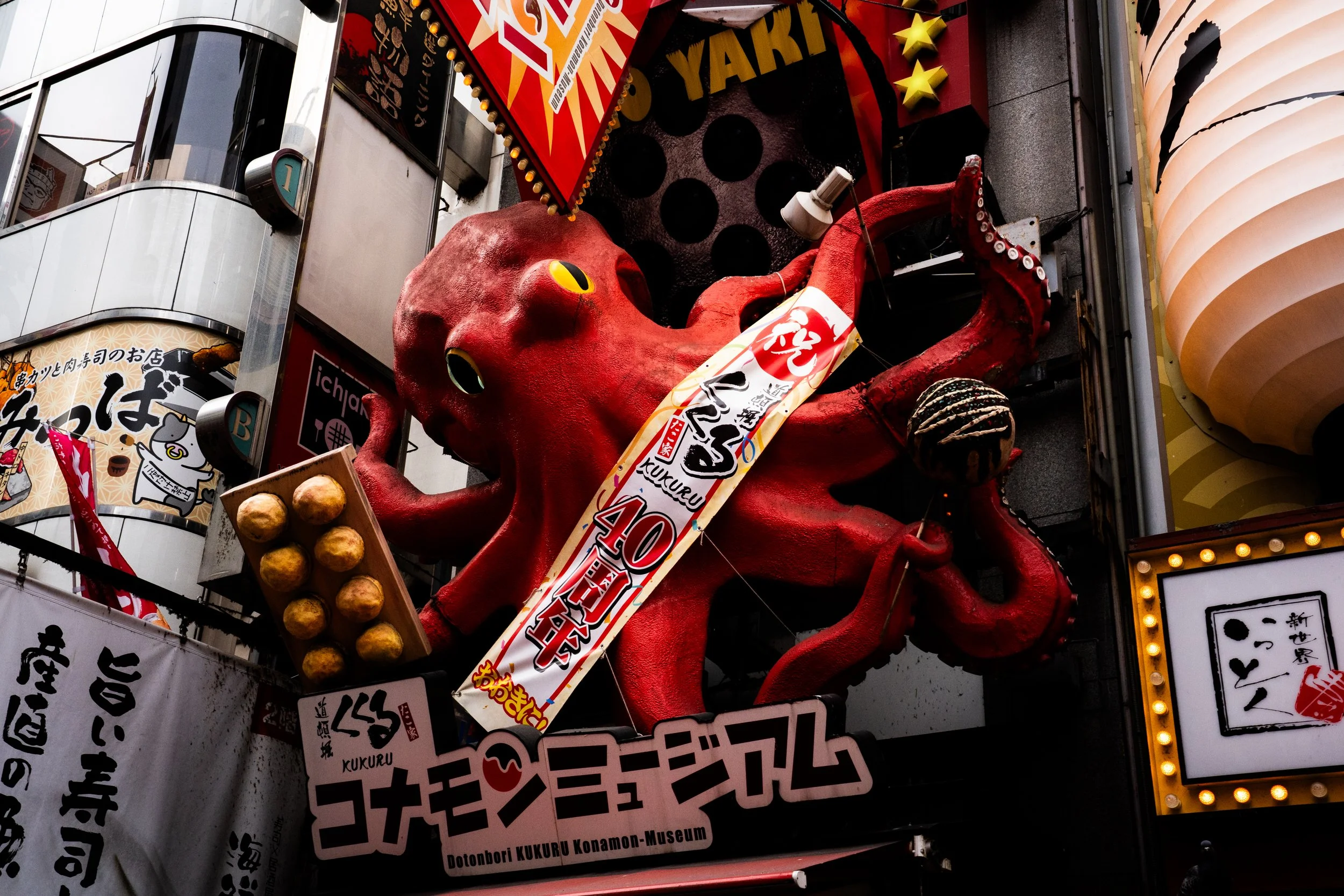 A large red octopus sculpture with yellow eyes, holding what appears to be a tray of fried foods, is surrounded by bright lights and signage in an Asian city street. The scene features multiple Japanese signs and advertisements.