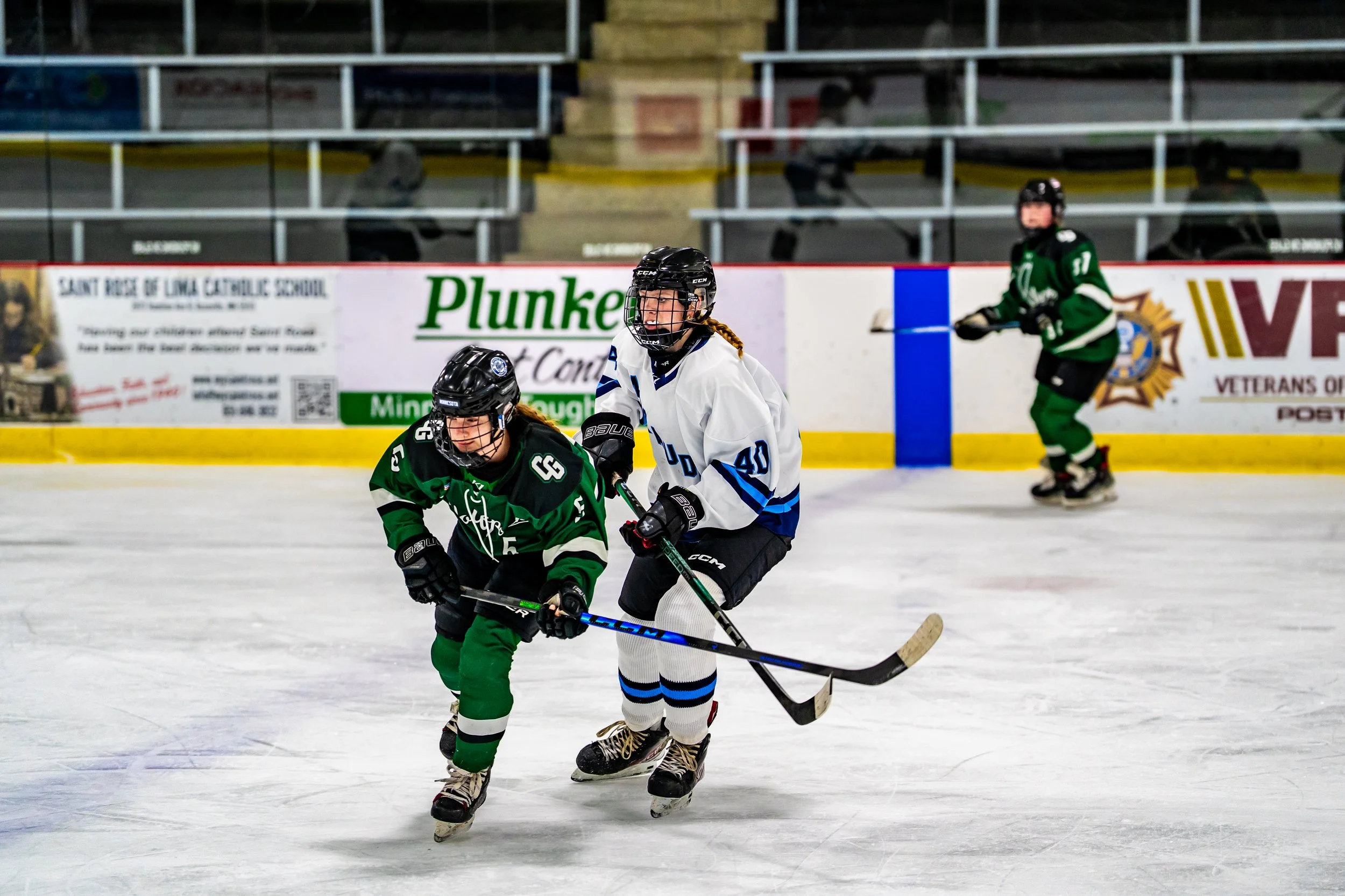 Three youth hockey players in gear on the ice rink, with two in green jerseys and one in a white jersey, competing for puck control during a game.
