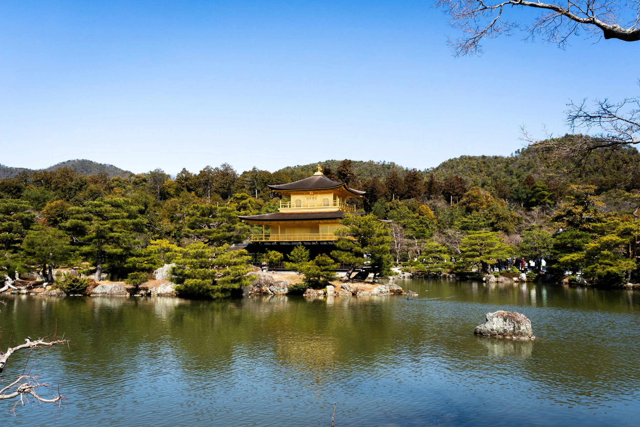 A traditional Japanese building, possibly a temple, situated among lush green trees by a body of water, with mountains in the background under a clear blue sky.