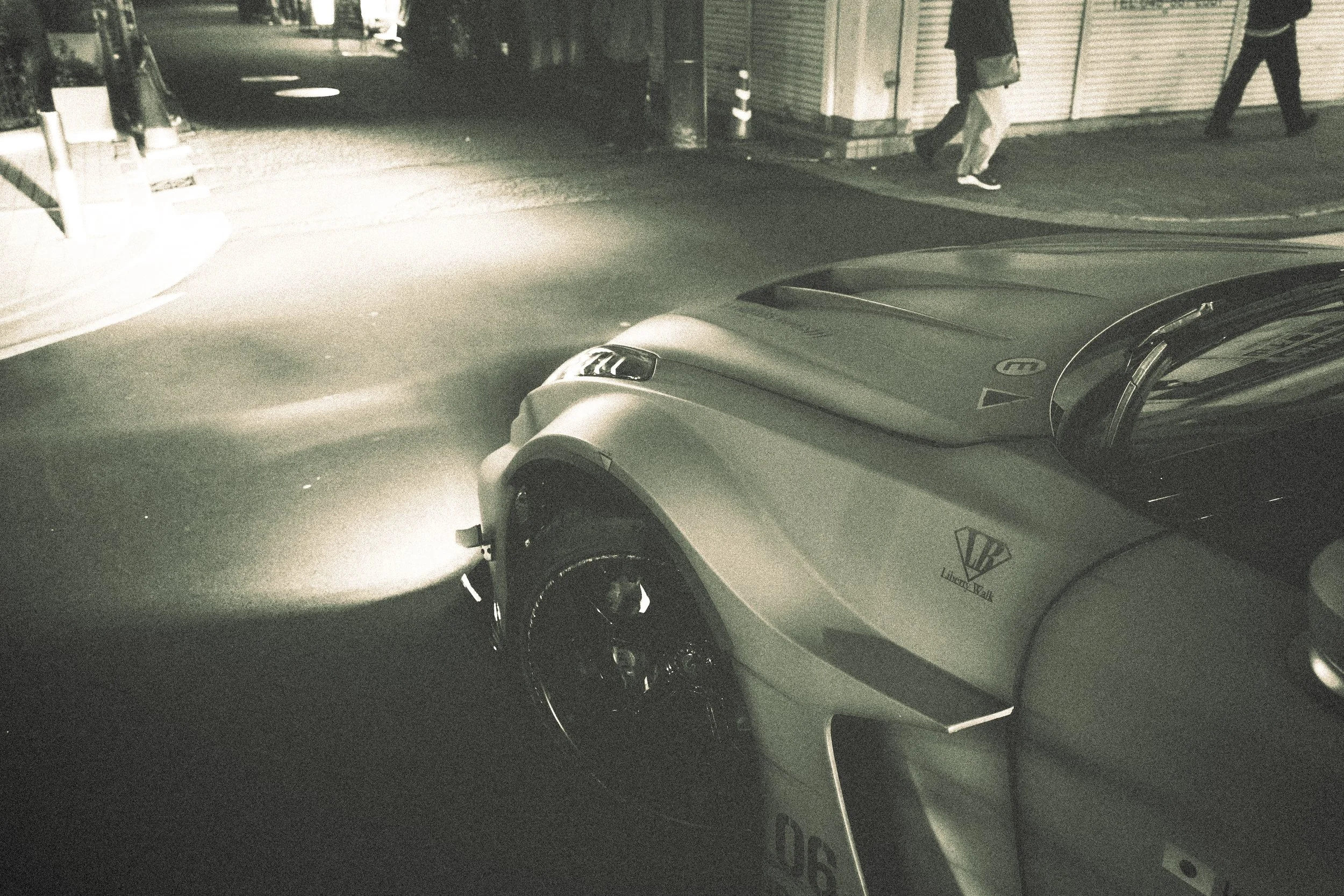 Nighttime street scene with a silver sports car in the foreground, two pedestrians walking past storefronts with closed shutters in the background, and a large dark object blocking part of the street.