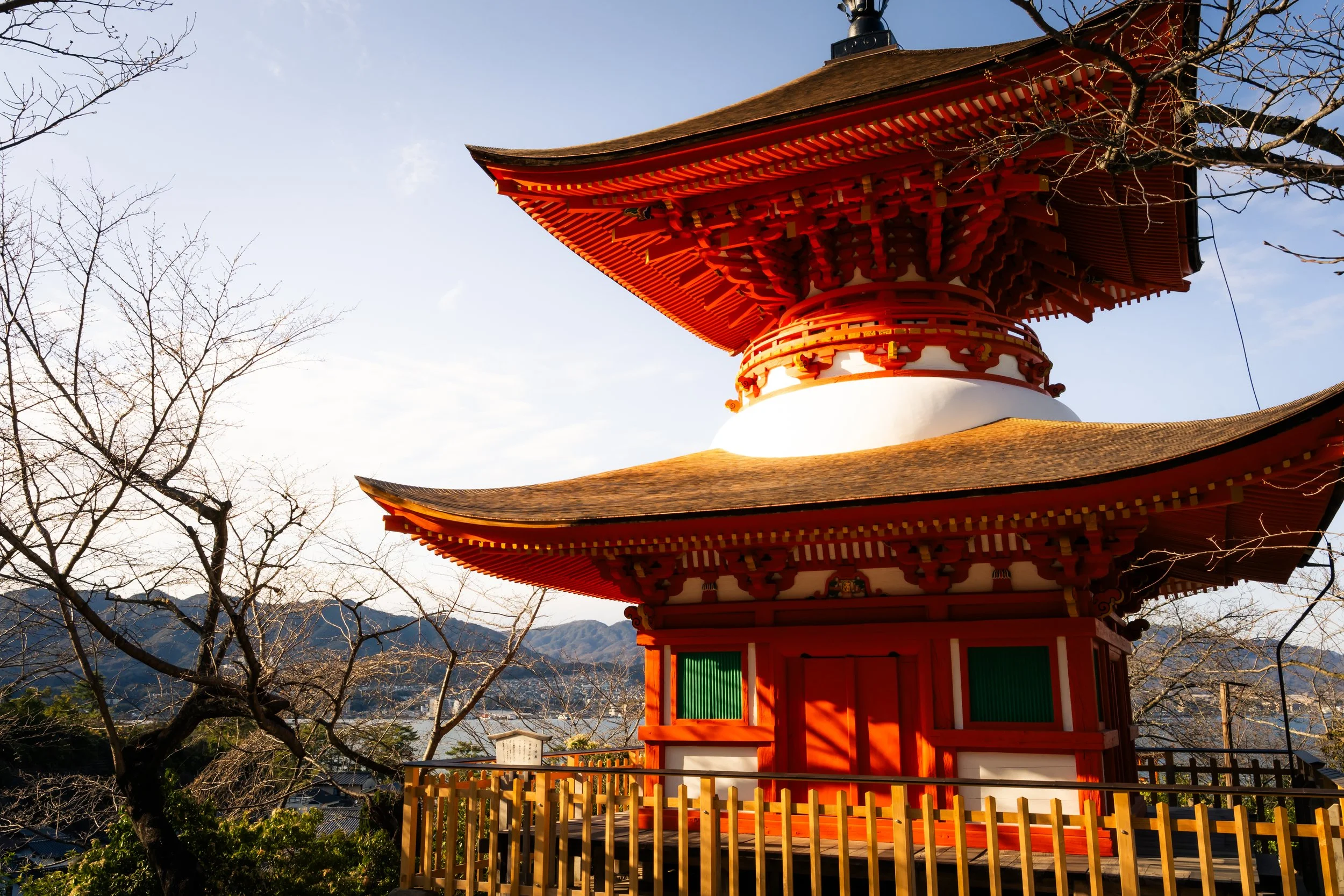 A traditional Japanese pagoda with multiple tiers, painted red and white, with a wooden fence in front and leafless trees around it, under a clear sky with mountains in the background.