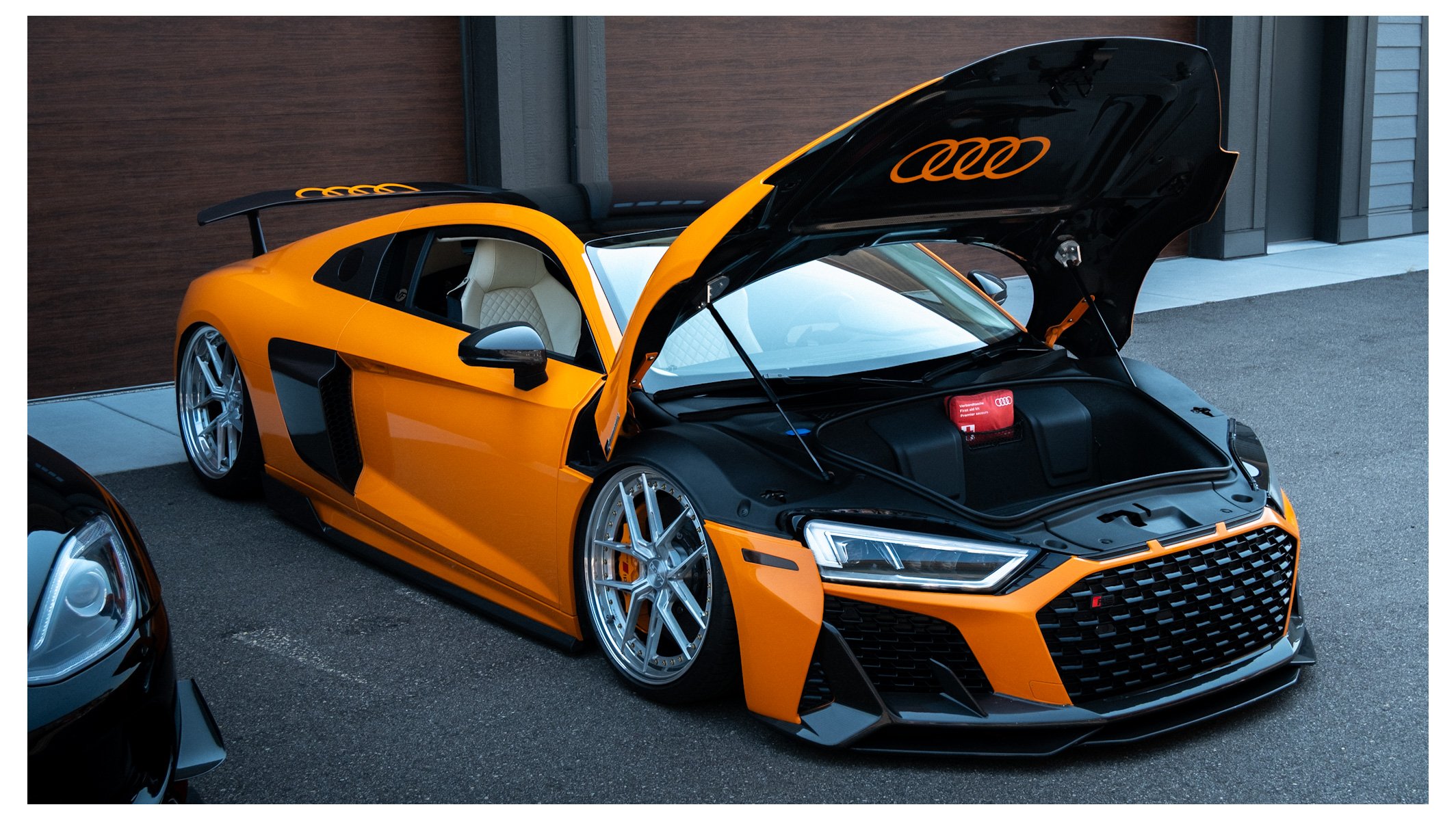 An orange Audi sports car with its hood open, parked in a driveway next to a wooden garage door and a gray building. The car has white interior seats and aftermarket silver wheels.