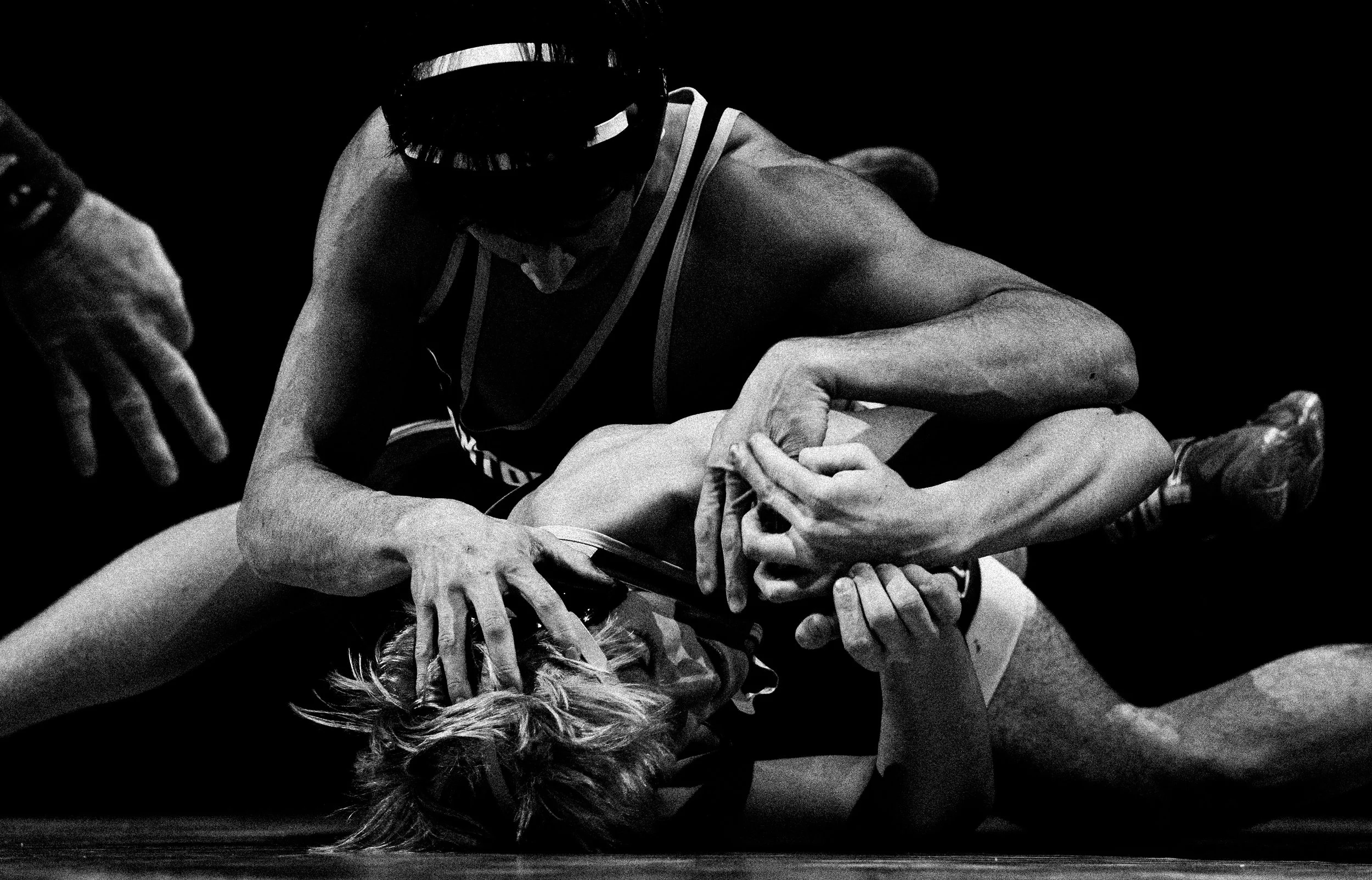 Black and white photo of two individuals engaged in a wrestling match, with one on top gripping the other's arm, the scene is intense and focused.