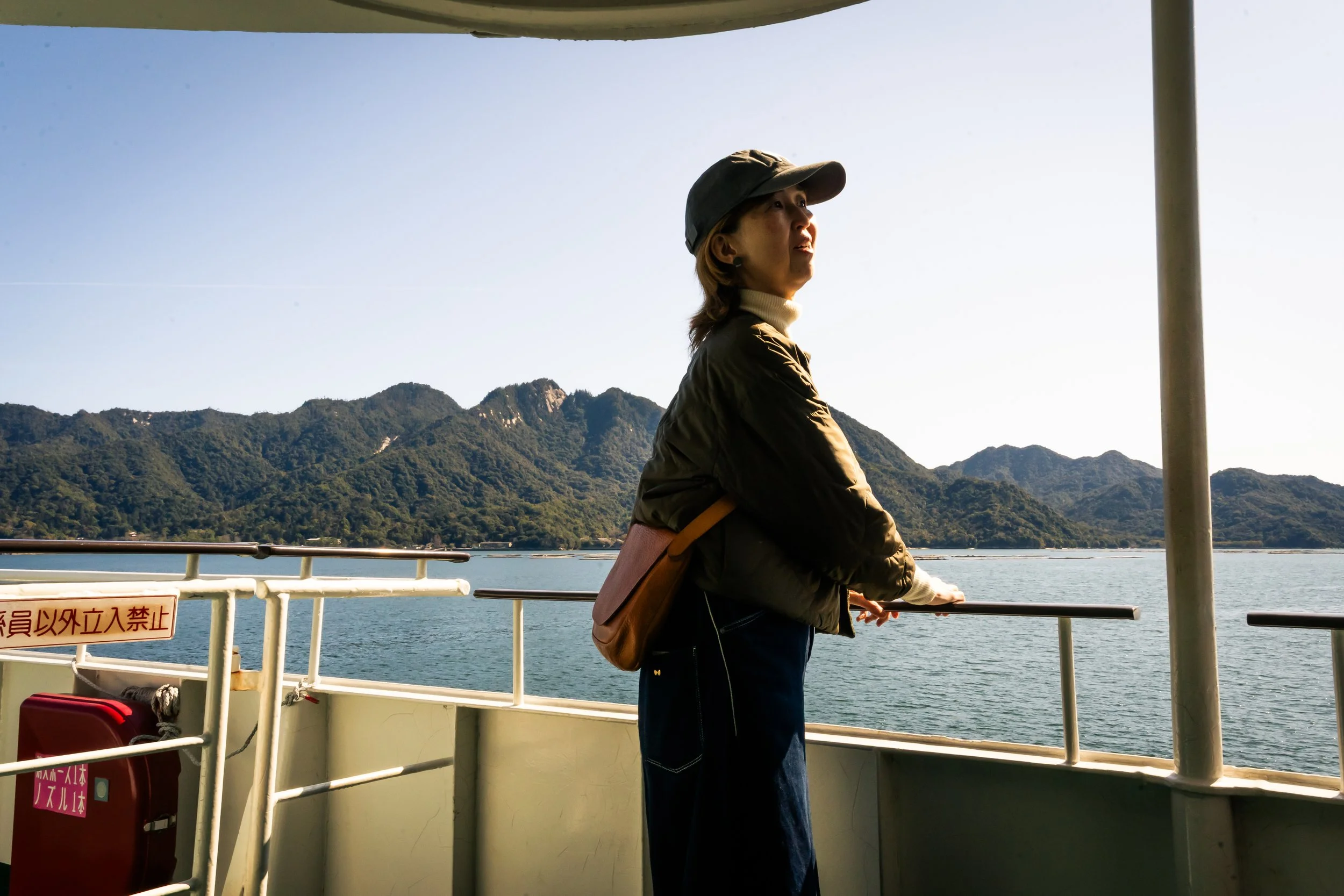 Woman wearing a hat and jacket standing on a boat with mountains in the background and water below.