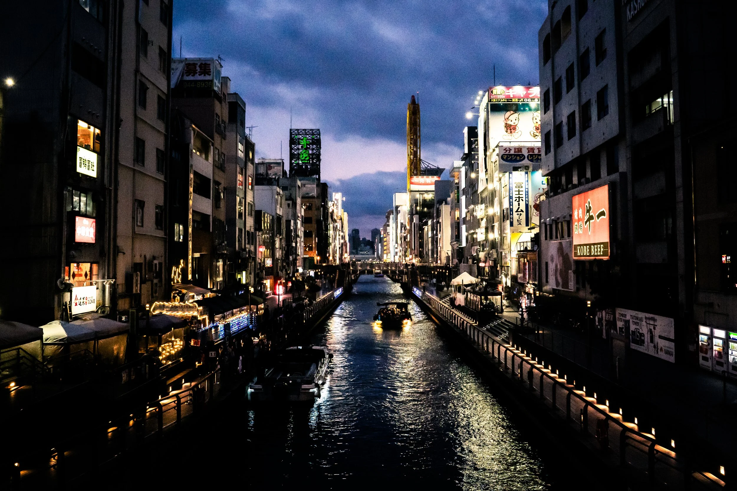 Nighttime cityscape with illuminated buildings and signs along a canal with boats, under a cloudy sky.