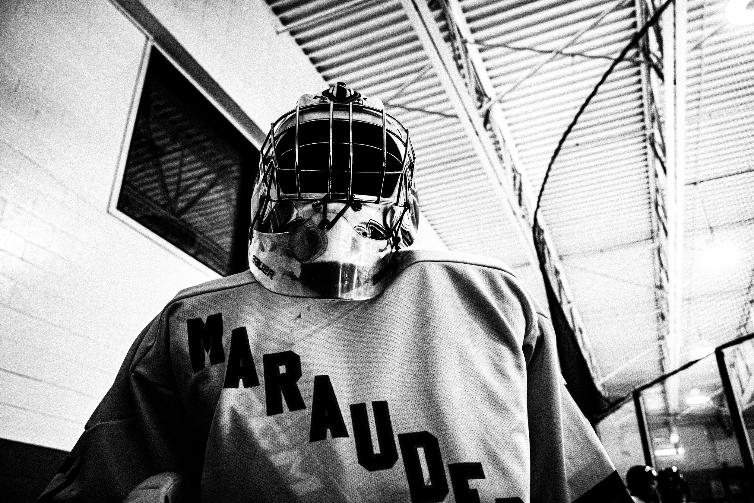 Close-up of an ice hockey goalie wearing a helmet with a face cage and a jersey with text, standing in front of an indoor arena with metal beams and structural elements overhead.
