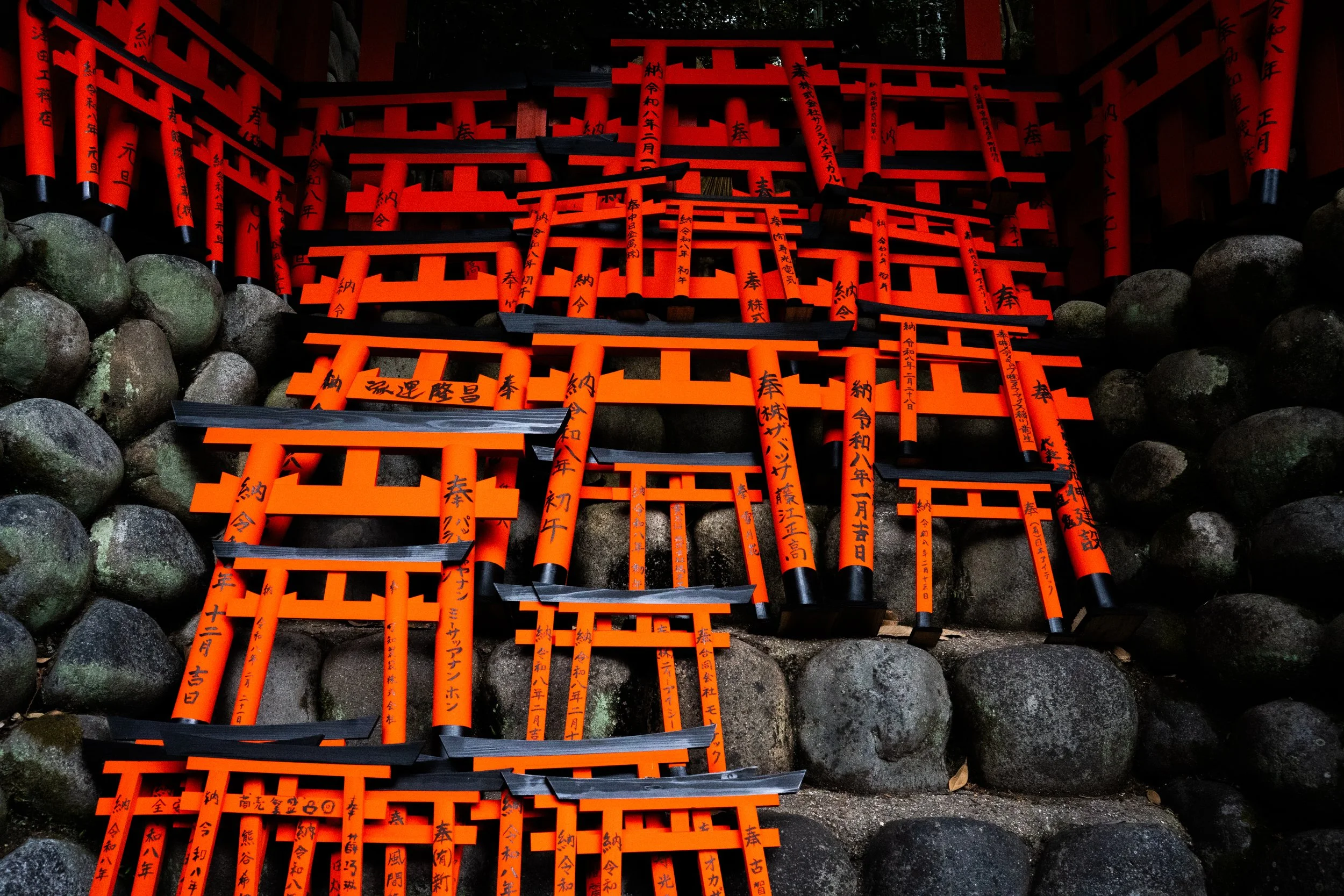A collection of small orange torii gates placed on stones at a Japanese shrine, with handwritten black characters on each gate.