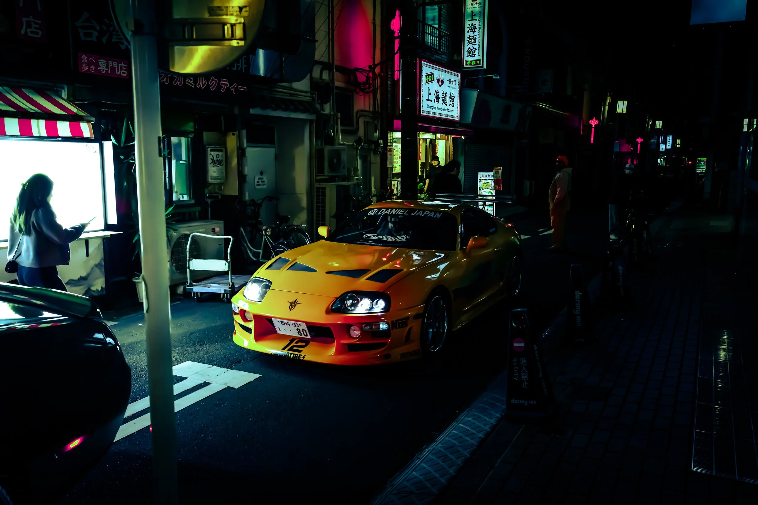 A brightly colored yellow sports car with racing decals parked on a city street at night. Neon signs with Asian characters light up the background, and several pedestrians are visible.