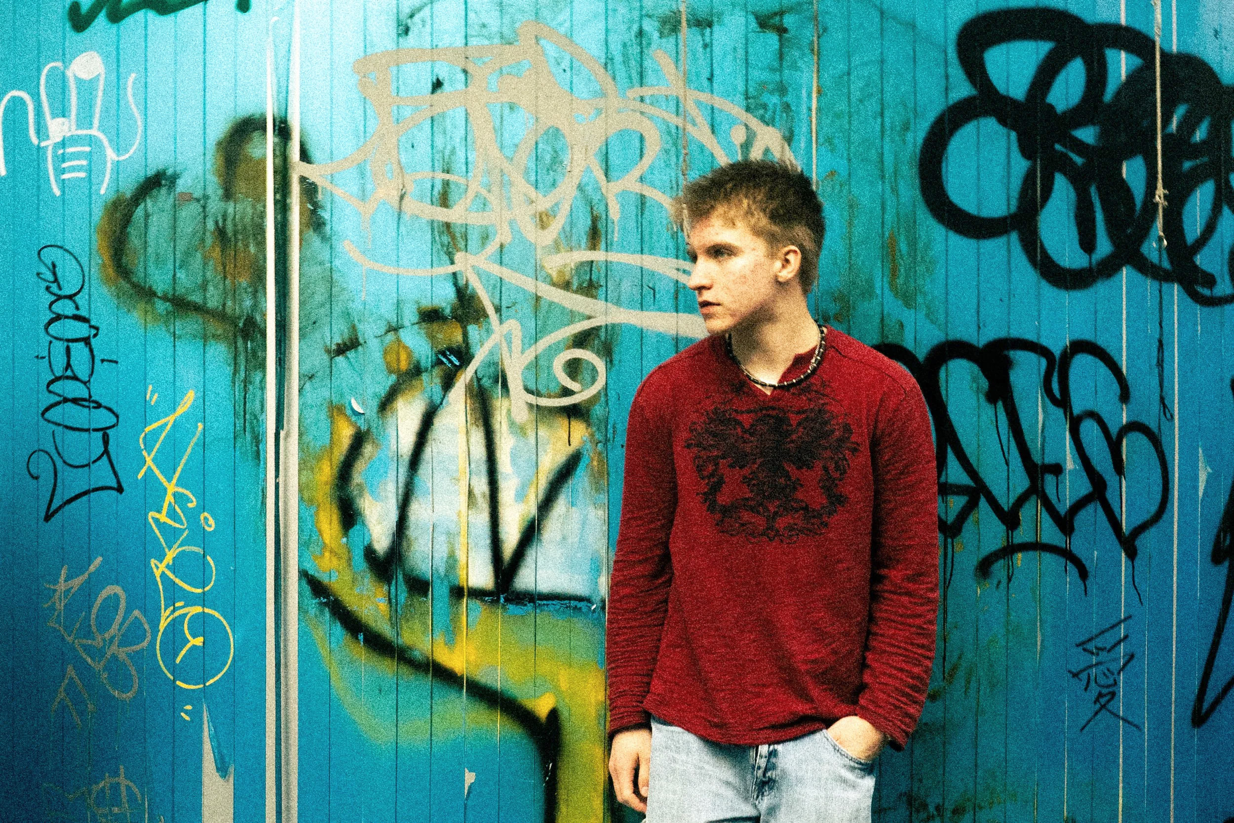 A young man with short hair, wearing a red long-sleeve shirt with a black graphic, light blue jeans, and a beaded necklace, standing against a graffiti-covered blue wooden wall.