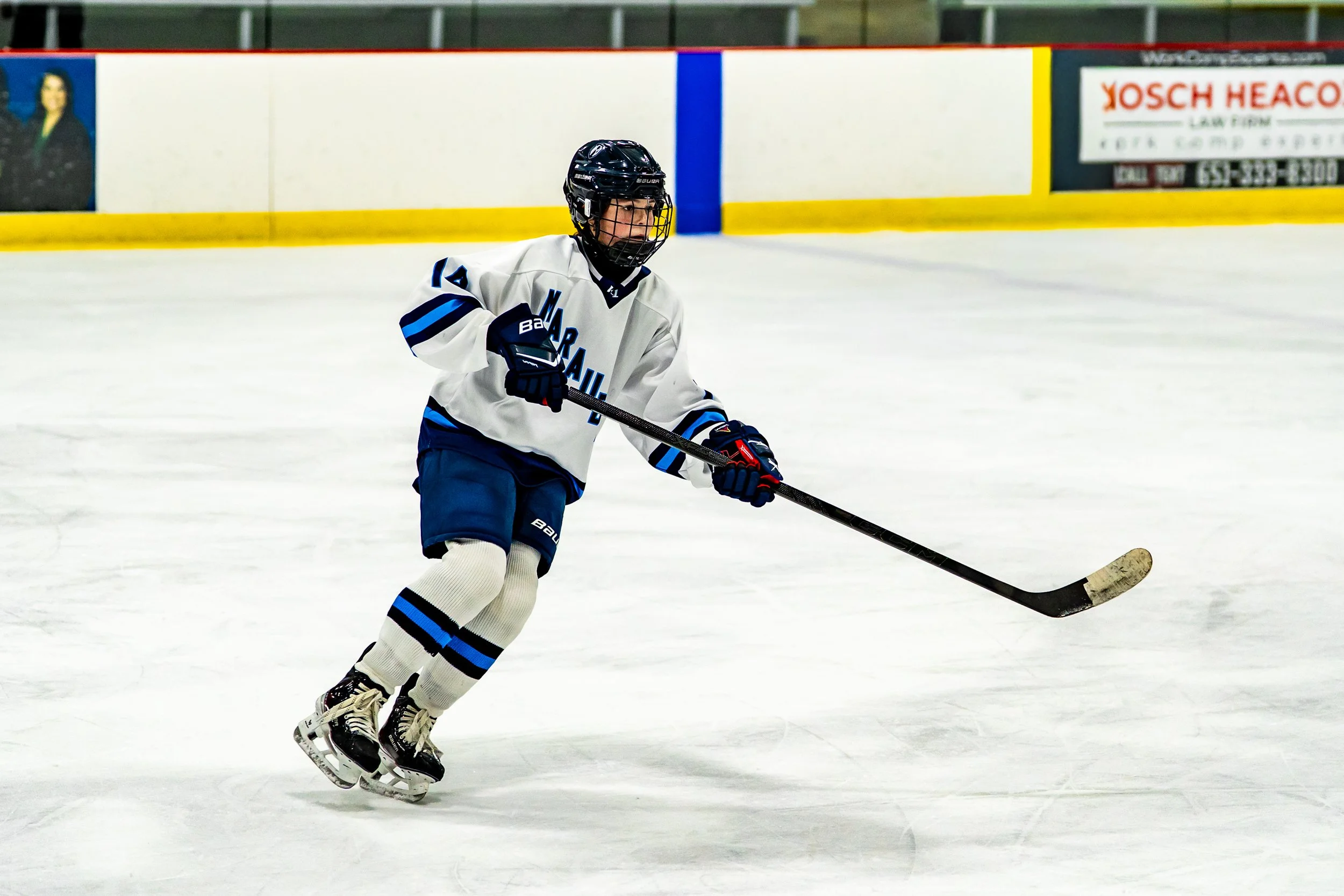 A young ice hockey player in a white and blue uniform skating on the ice with a hockey stick.