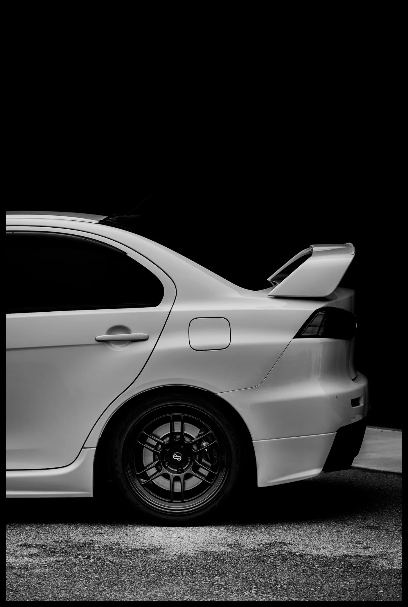 A black and white photo of the rear side of a white car with a distinctive spoiler on the trunk, parked on a textured asphalt surface.