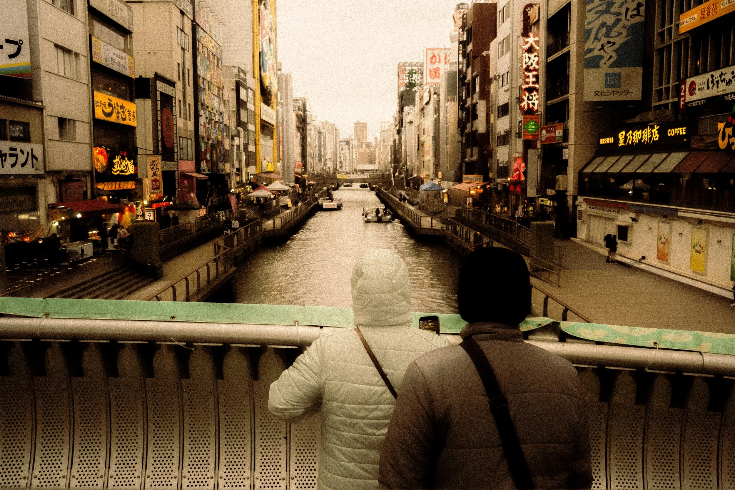 Two people sitting in front of a window overlooking a city canal with buildings and neon signs.