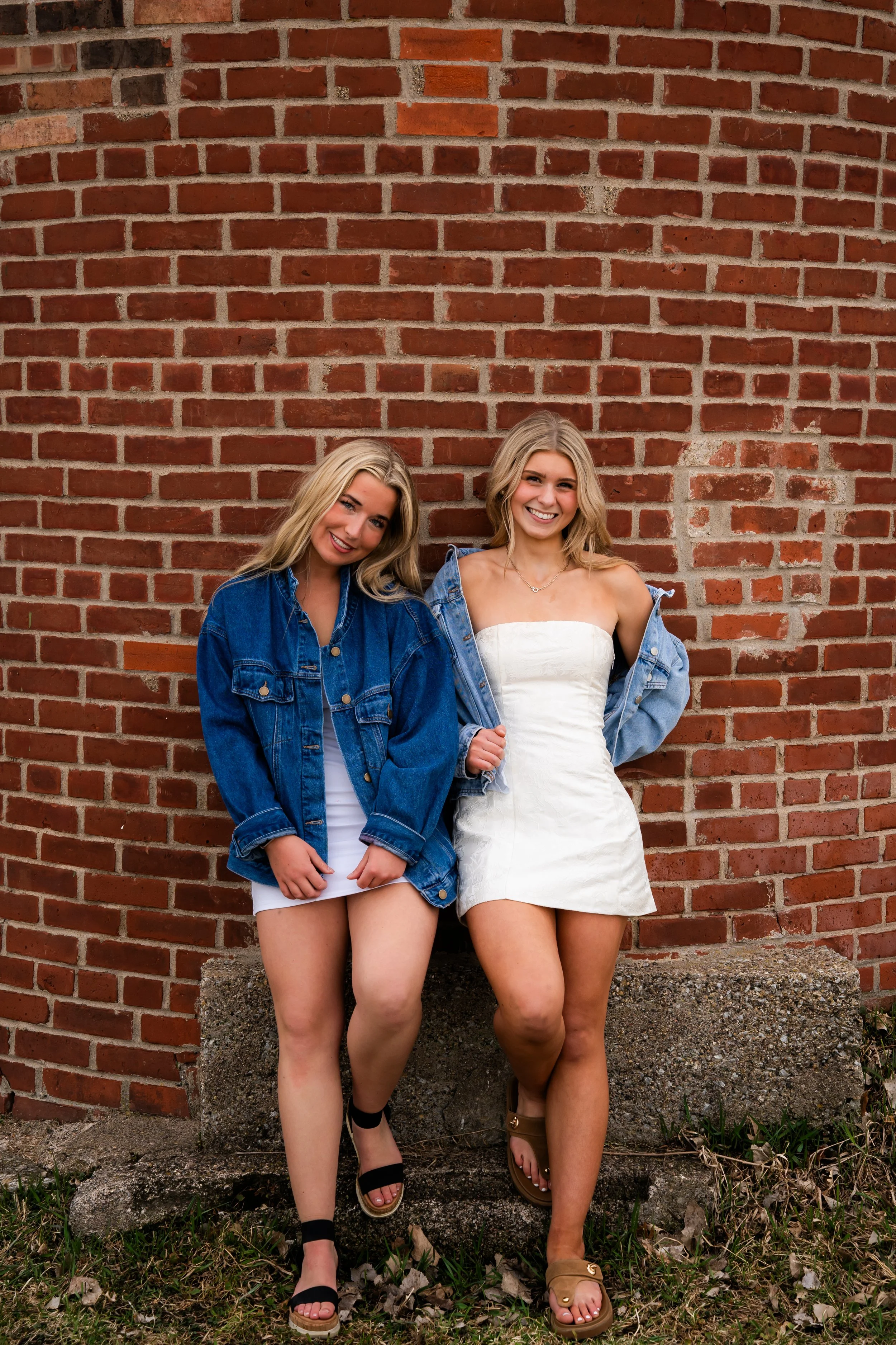 Two young women with blonde hair smiling and standing against a brick wall, wearing denim jackets and white dresses.