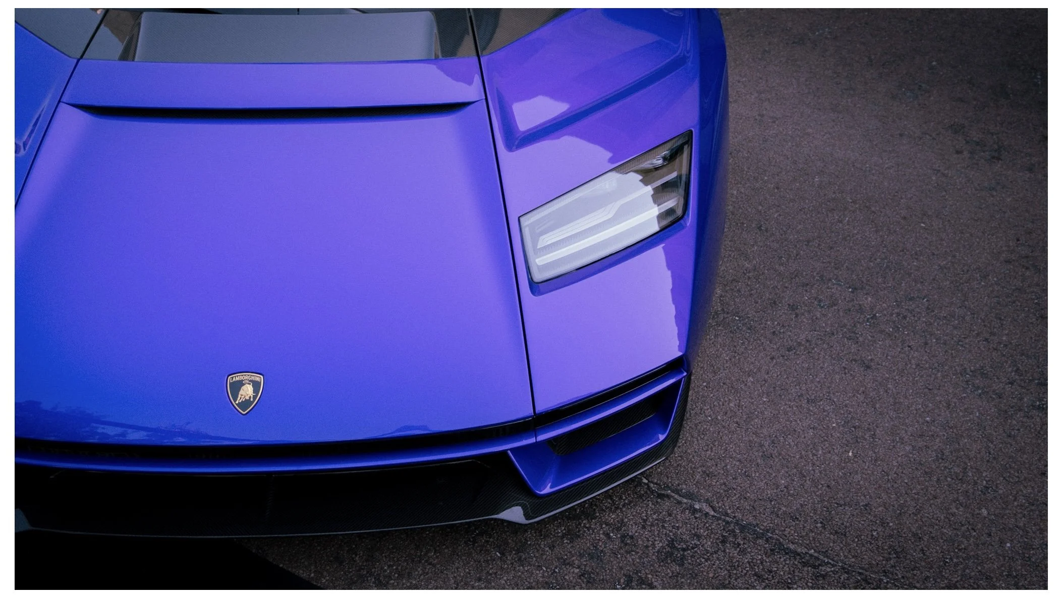 Close-up of the front part of a blue Lamborghini sports car, showing the Lamborghini emblem on the hood and part of the headlight and front bumper, on a paved surface.