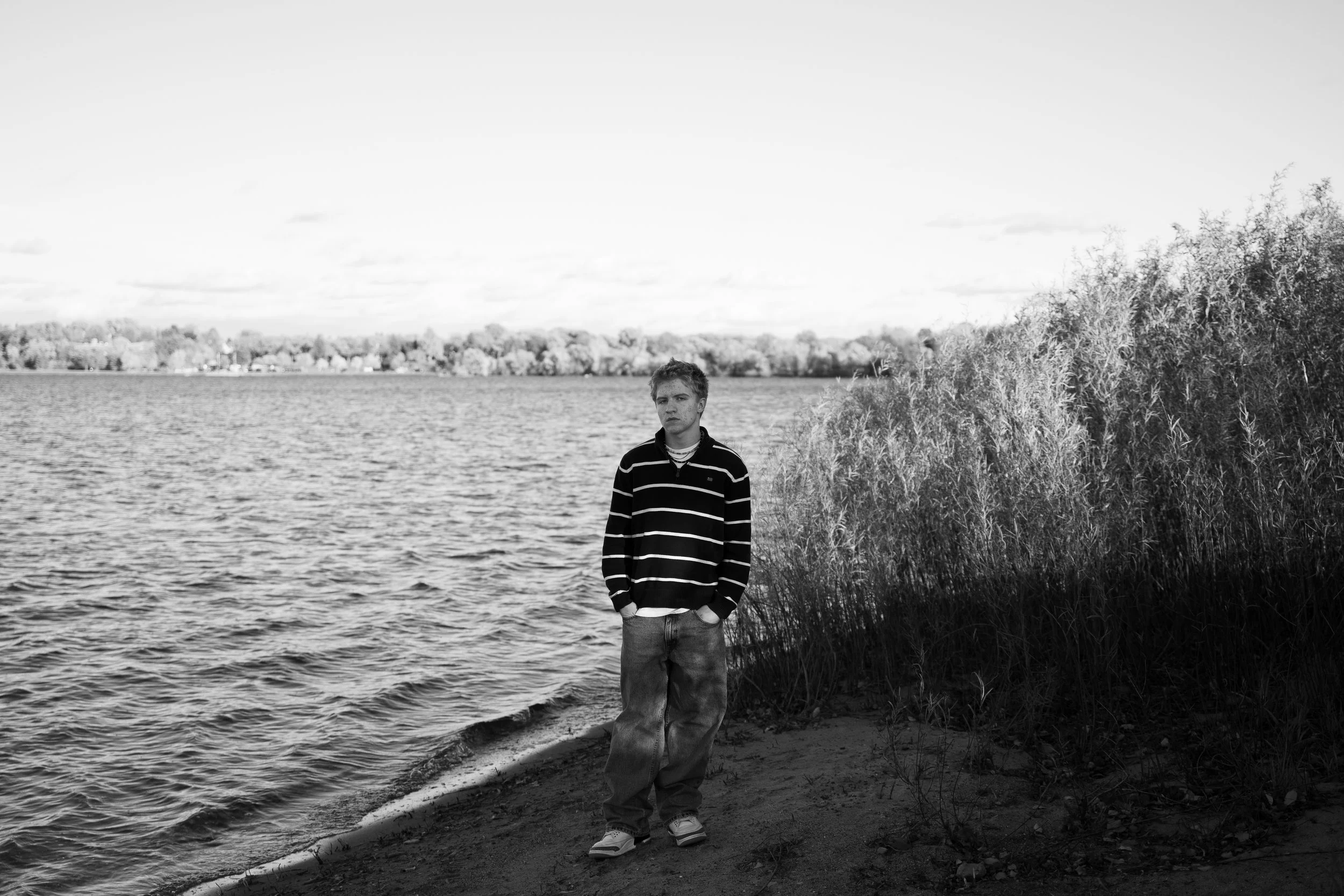 A young man stands on the shoreline of a lake or river, with trees and a distant town or city in the background. He is wearing a striped hoodie and jeans, and is looking slightly to the side with a neutral expression.