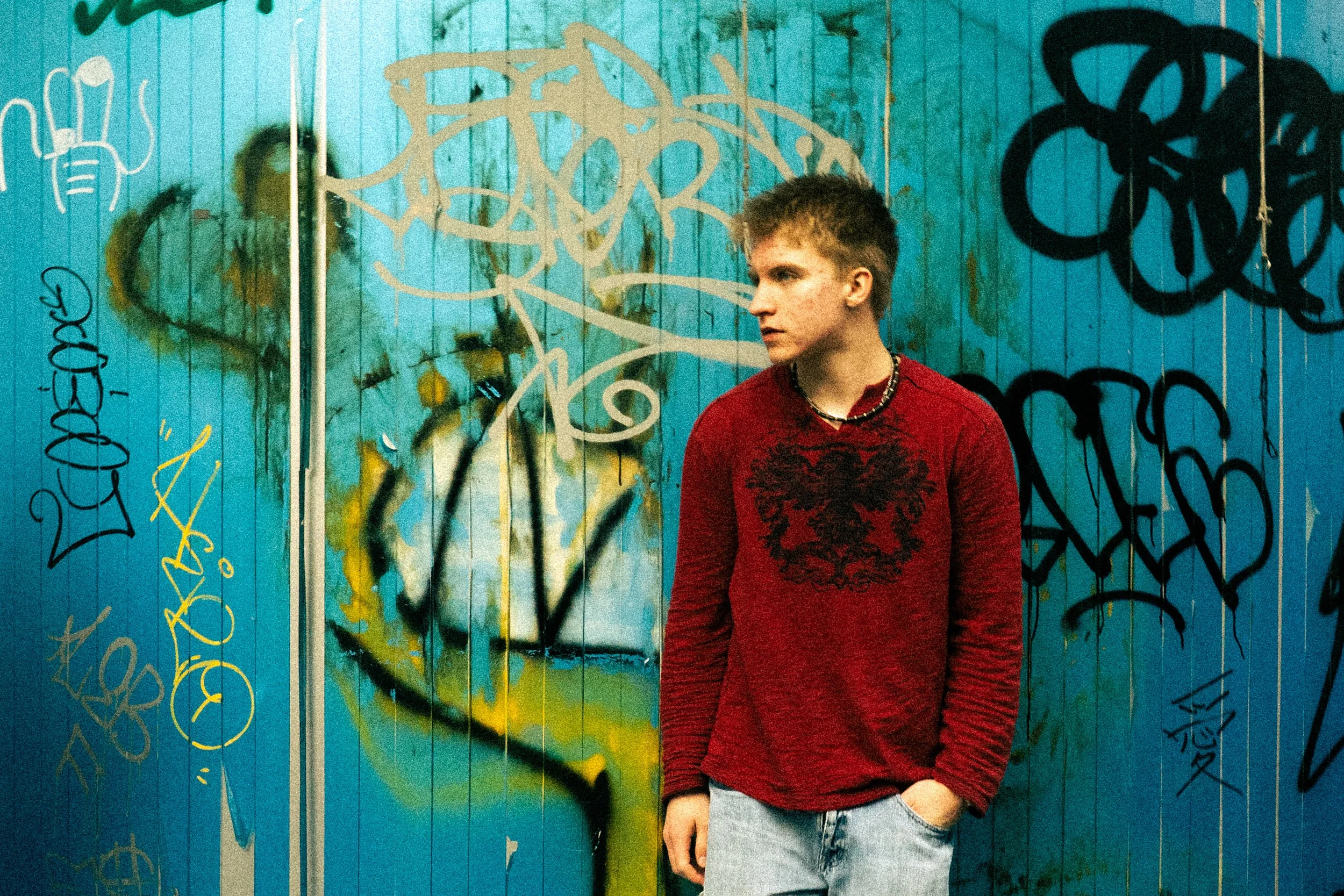 A young man with spiky hair wearing a red long-sleeve shirt with a black graphic and gray shorts standing against a vibrant blue graffiti-covered wall.