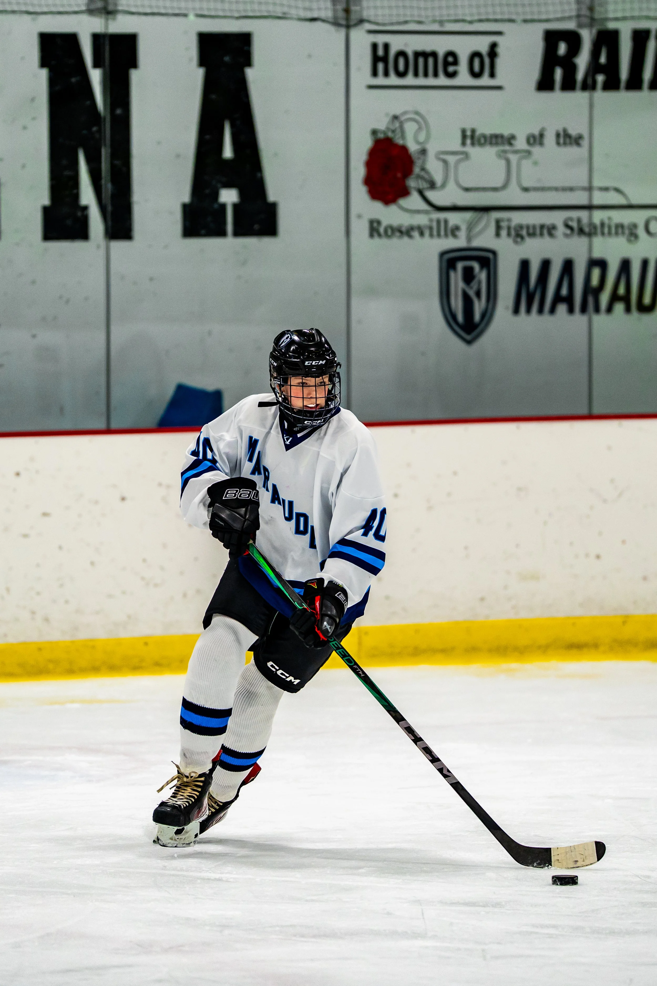 A young hockey player skating on the ice rink, wearing a white jersey with blue and black accents, a black helmet, and holding a hockey stick, with a black puck in front of them.