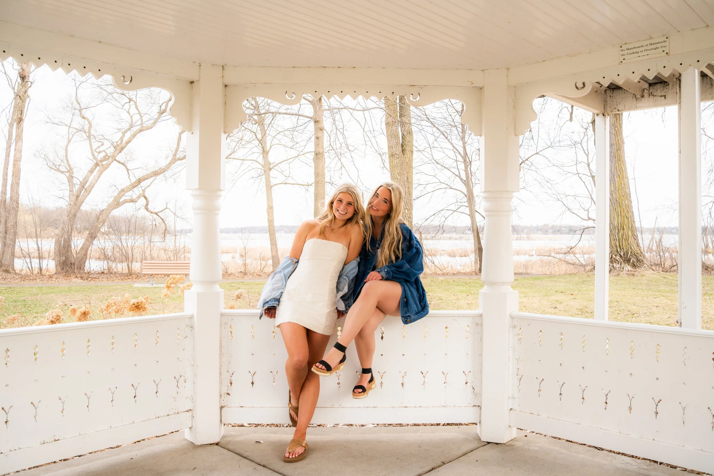 Two young women sitting together on a white gazebo with a park and leafless trees in the background. One woman is wearing a white dress and the other is wearing a blue jacket. They are smiling, embracing, and appear to be enjoying a casual outdoor mo