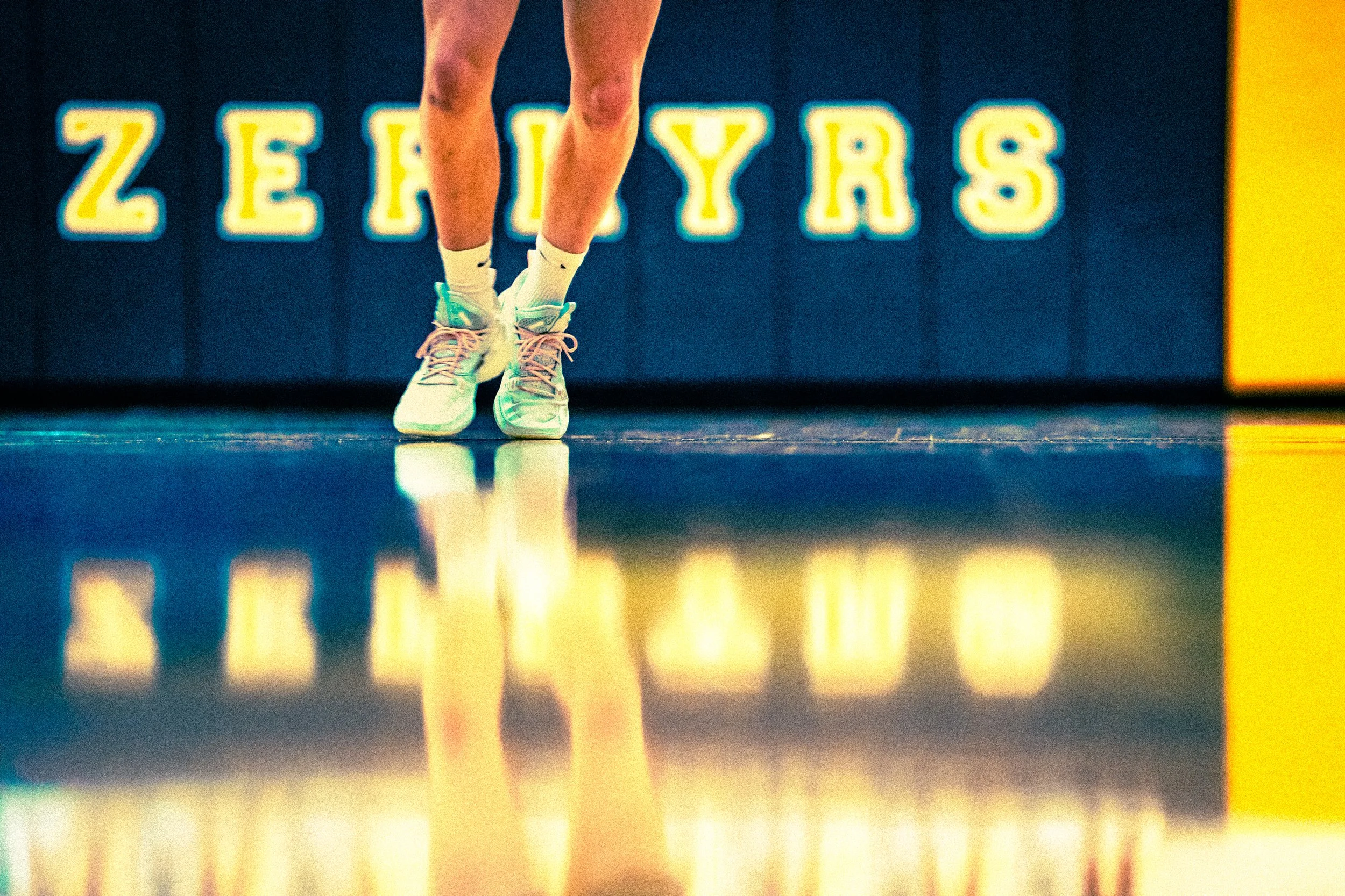 Close-up of a runner's legs and feet wearing white and teal athletic shoes, running on an indoor track with yellow and blue signage in the background.