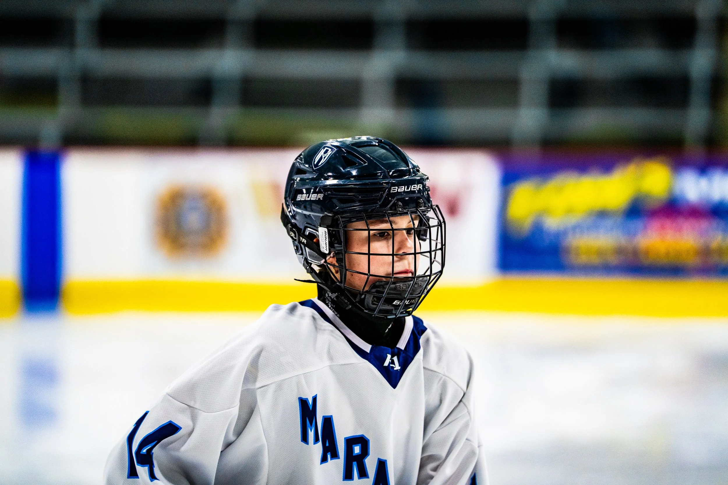 An ice hockey player wearing a black helmet and a white jersey with blue lettering, on an ice rink. The background features the ice rink boards and out-of-focus advertisements.